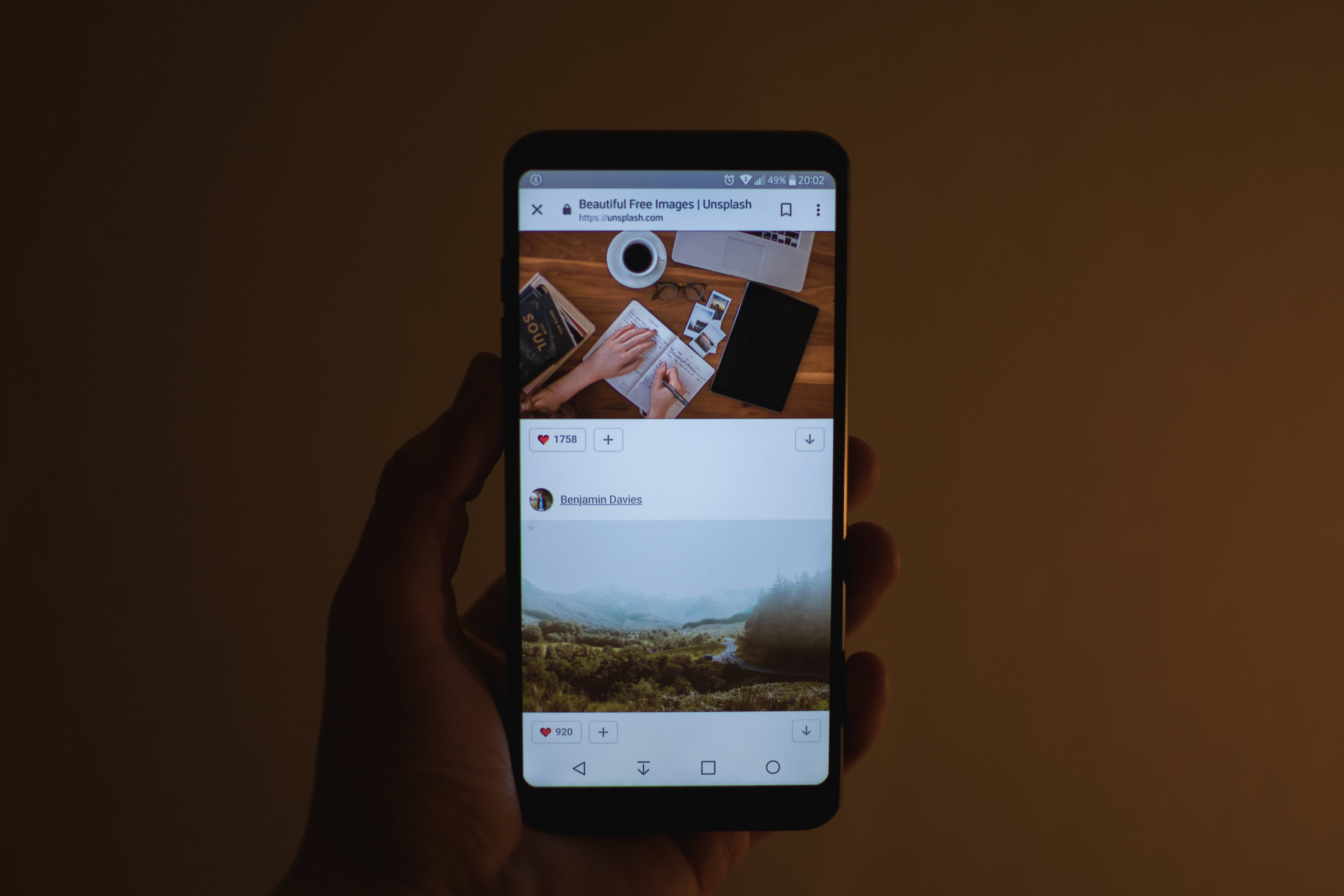 Lg 桌布 壁紙 — 一隻手拿著智慧型手機，顯示 Unsplash 網頁，上面有山景照片和木桌上的一杯咖啡。