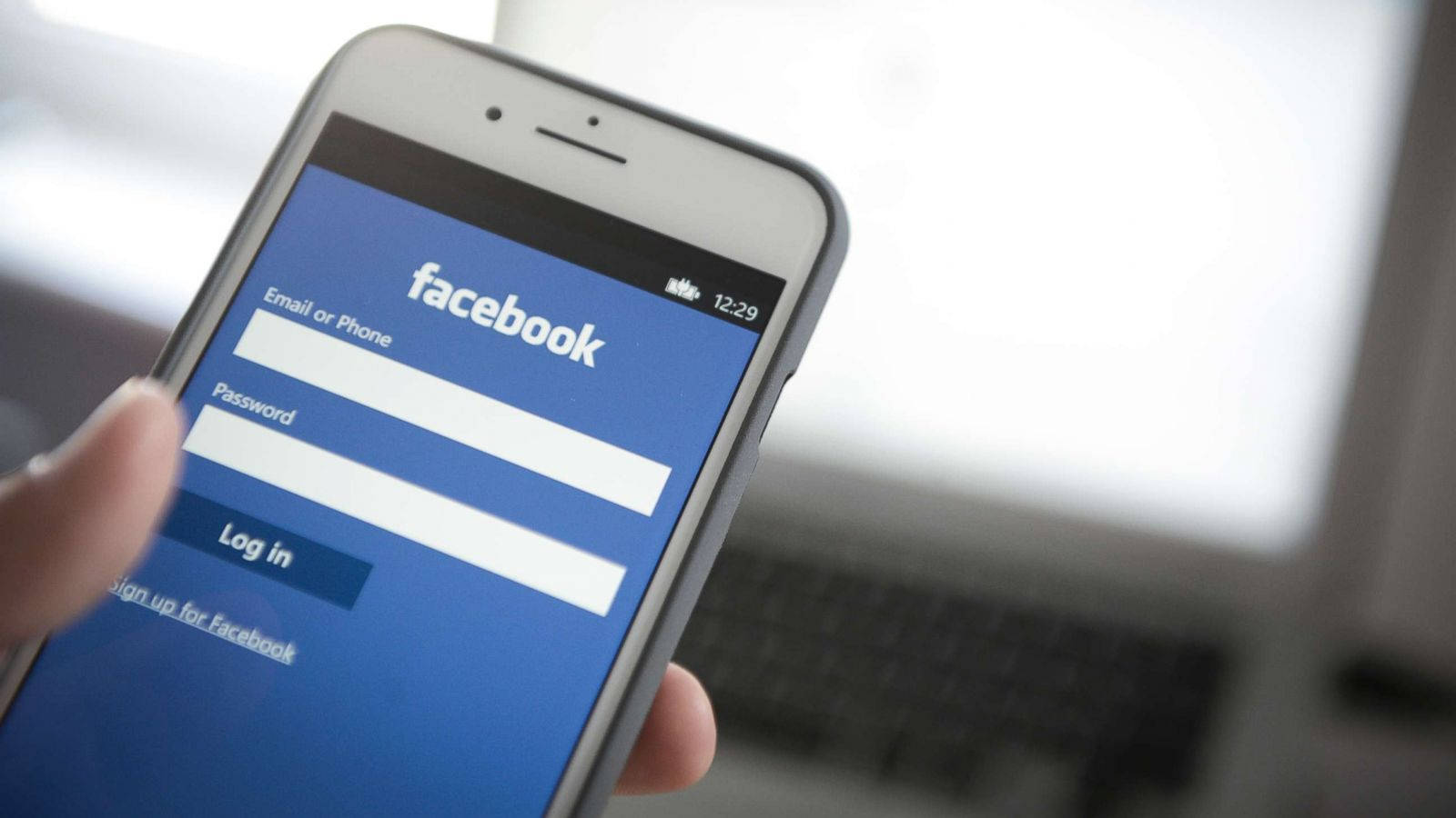 Facebook Login 桌布 壁紙 — 一隻手拿著智慧型手機，顯示Facebook登入畫面，背景有模糊的筆記型電腦。