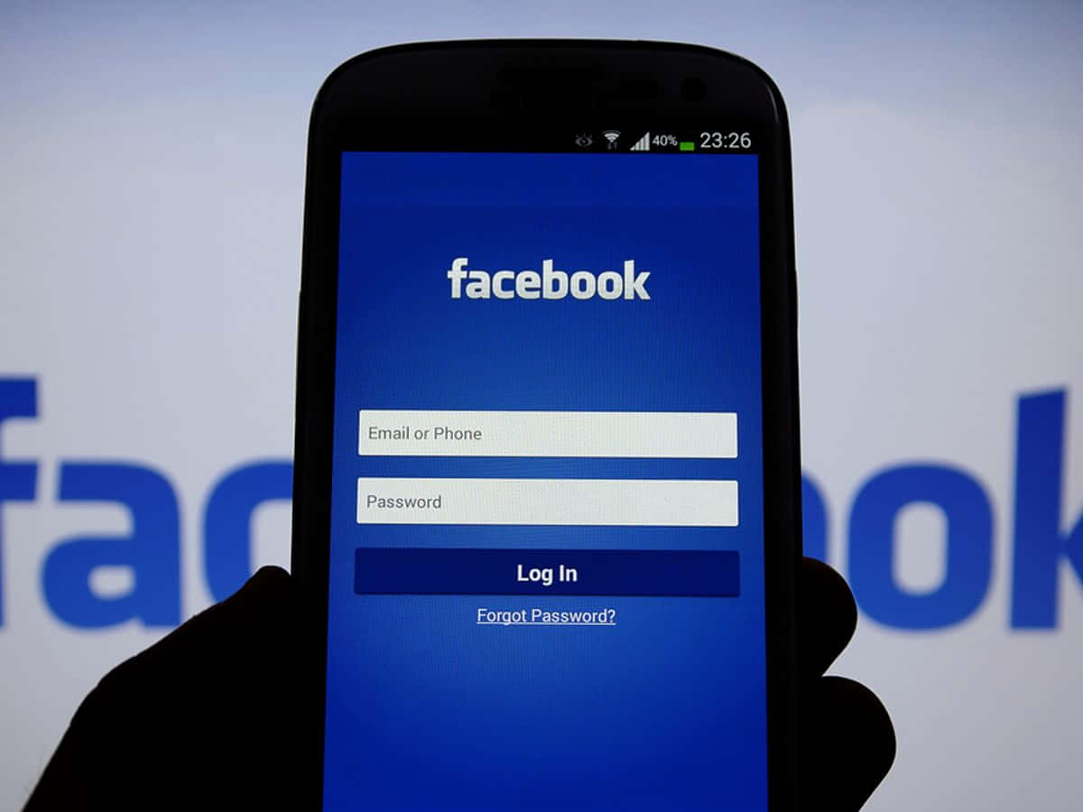Facebook 桌布 壁紙 — 一隻手拿著智慧型手機，顯示Facebook登入畫面，背景是模糊的Facebook標誌。