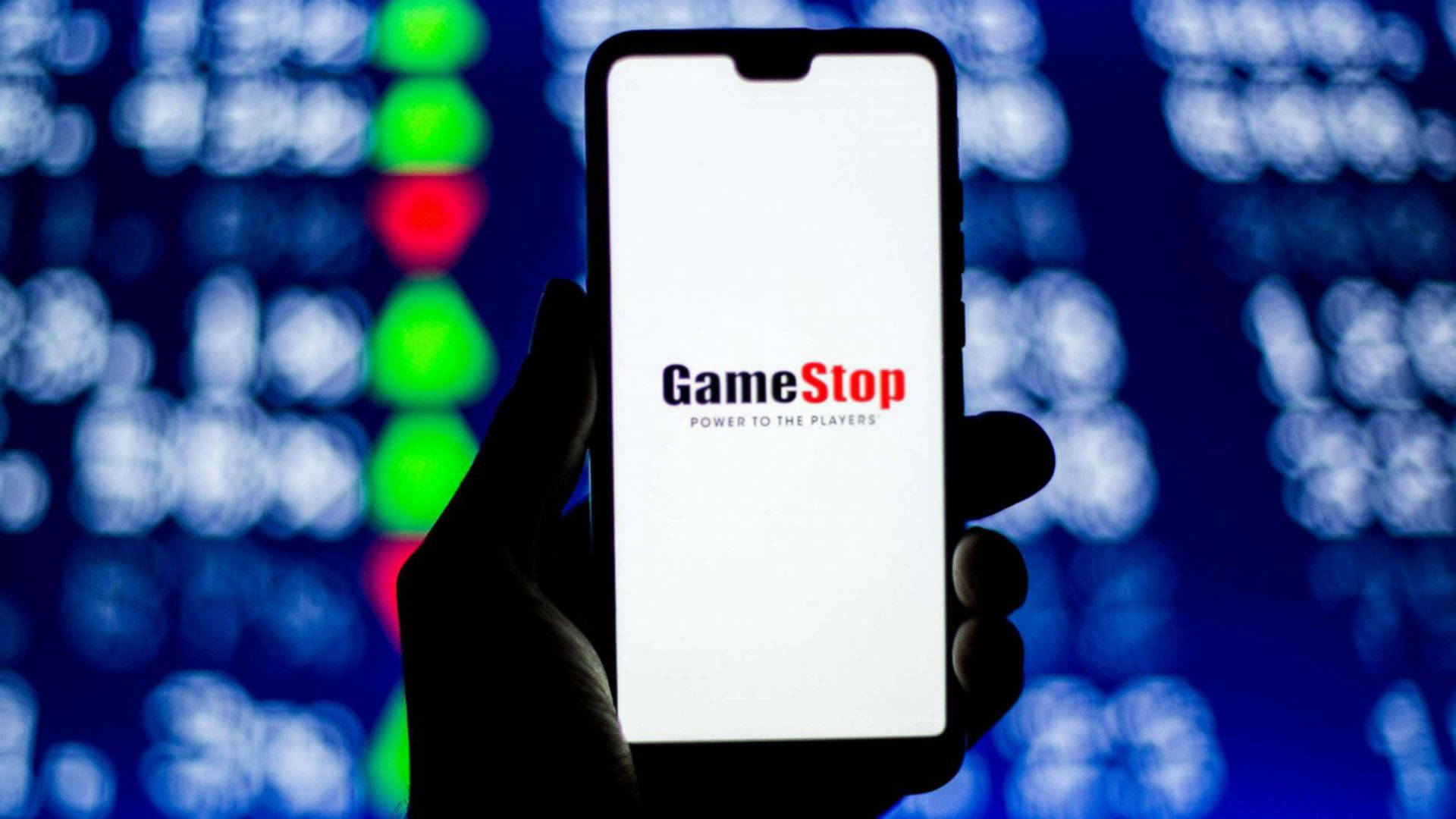 GameStop 桌布 壁紙 — 一隻手拿著智慧型手機，顯示GameStop標誌，背景是模糊的股票市場行情燈光。