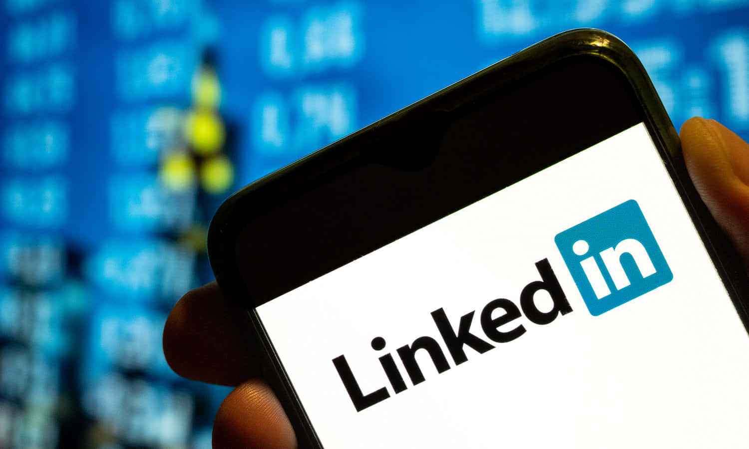 手機 桌布 壁紙 — 一隻手拿著智慧型手機，顯示LinkedIn標誌，背景是模糊的藍色股票報價板。