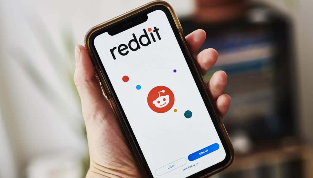 Reddit 桌布 壁紙 — 一隻手拿著智慧型手機，顯示Reddit應用程式的登入畫面，帶有其標誌性的外星人吉祥物和彩色圓點。