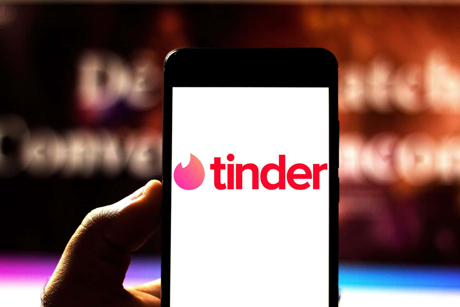 tinder 桌布 壁紙 — 一隻手拿著智慧型手機，顯示Tinder應用程式標誌，背景模糊色彩繽紛。