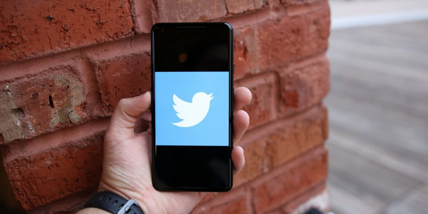 Twitter 桌布 壁紙 — 一隻手拿著智慧型手機，顯示Twitter標誌，背景是紅磚牆。