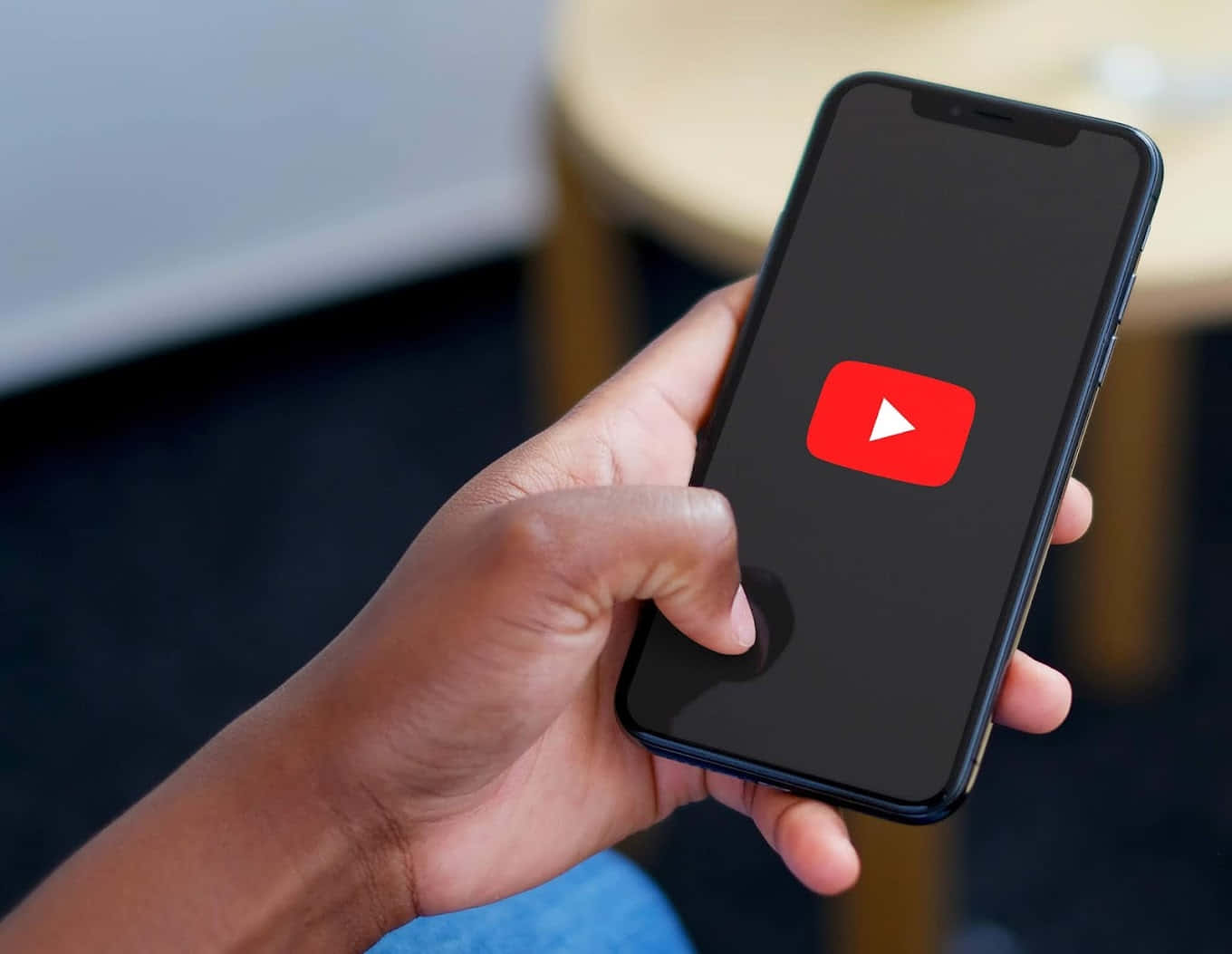 YouTube 桌布 壁紙 — 一隻手拿著智慧型手機，顯示YouTube應用程式圖示，背景為模糊的室內環境。