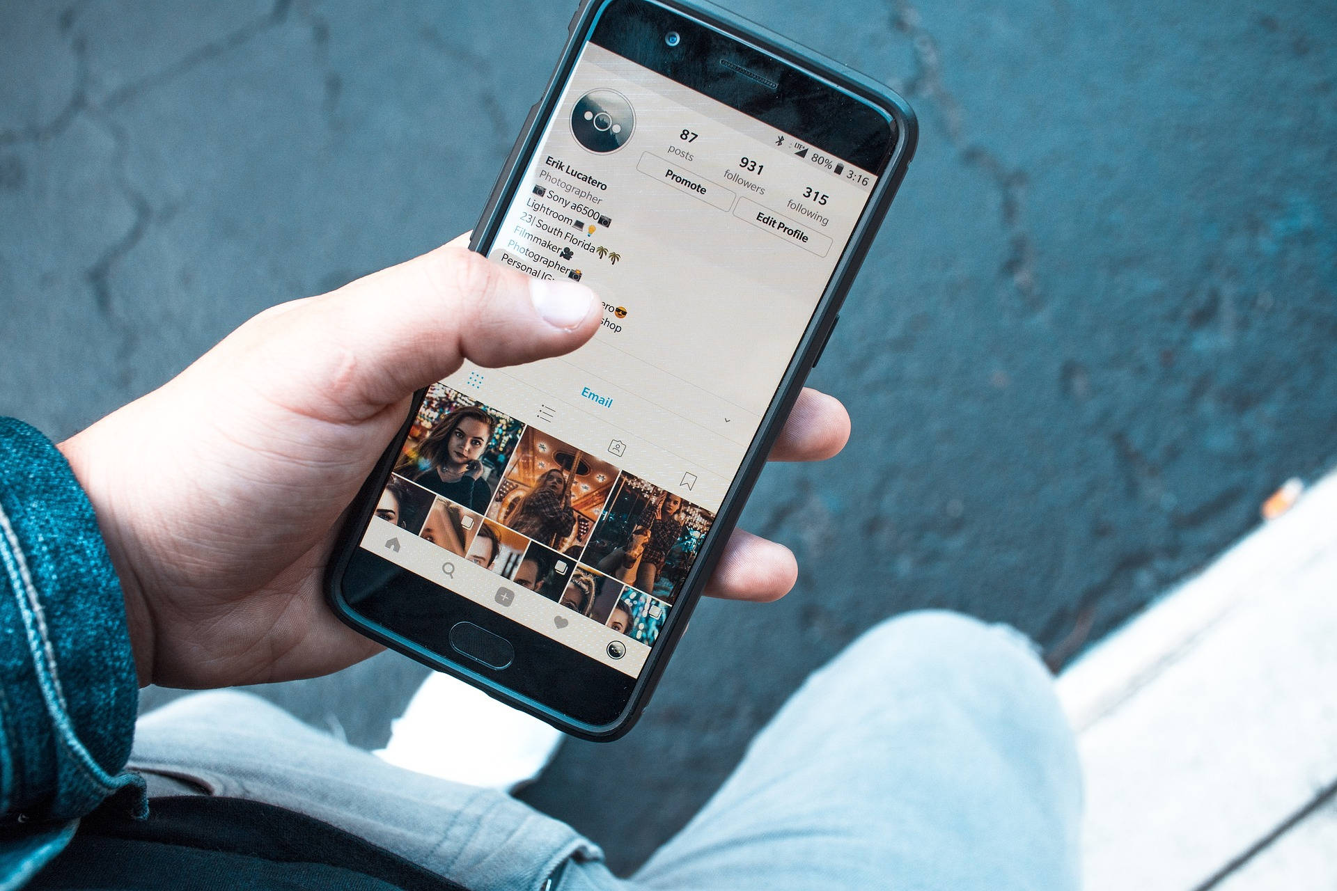Mobile Phone 桌布 壁紙 — 一隻手拿著智慧型手機，顯示一個Instagram個人資料，上面有照片網格，背景是紋理路面。