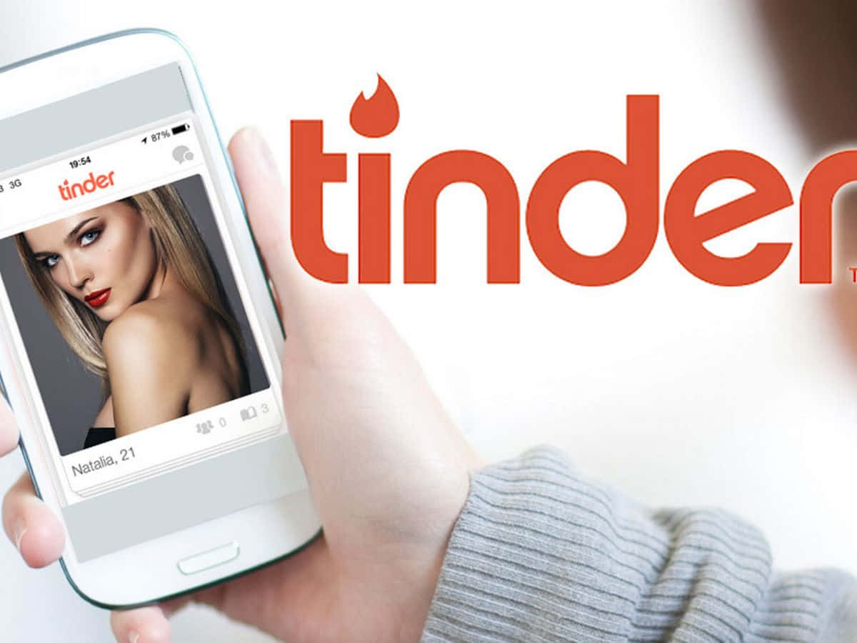 tinder 桌布 壁紙 — 一隻手拿著智慧型手機，顯示一個名為Natalia 21的女性的Tinder個人資料，背後有橙色的Tinder標誌。