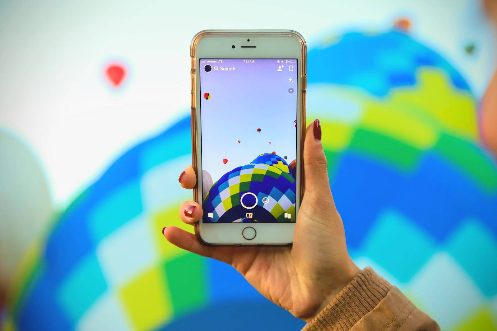 original iPhone 6 桌布 壁紙 — 一隻手拿著智慧型手機，顯示一個色彩繽紛的熱氣球應用程式，背景是充滿活力的抽象圖案。