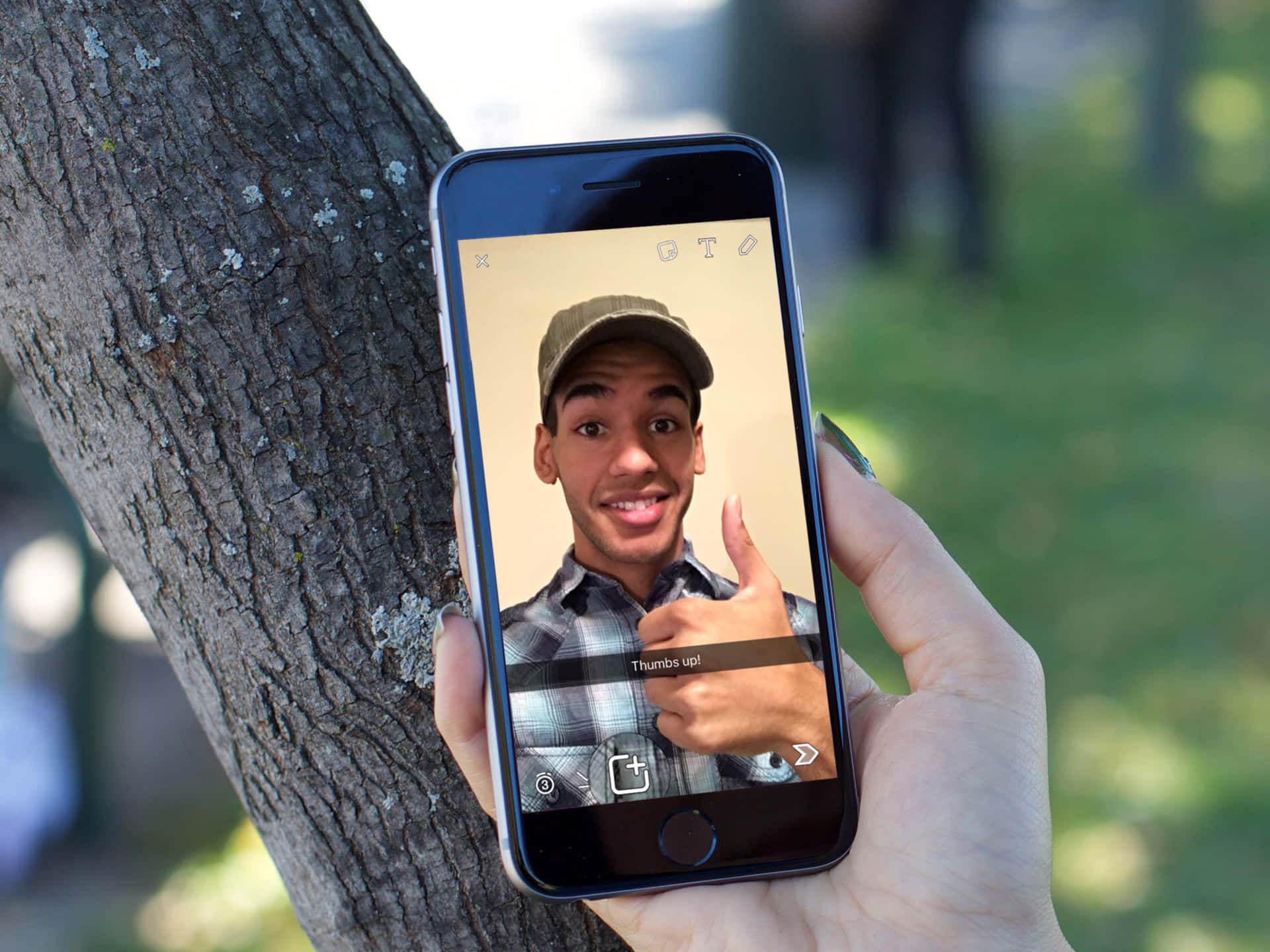 Snapchat 桌布 壁紙 — 一隻手拿著智慧型手機，顯示一個開心的年輕人在陽光明媚的公園裡靠著樹豎起大拇指。