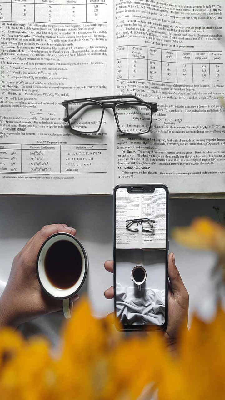 辦公室 桌布 壁紙 — 一隻手拿著智慧型手機，顯示一副眼鏡，鏡像出同一副放在化學教科書上的眼鏡。