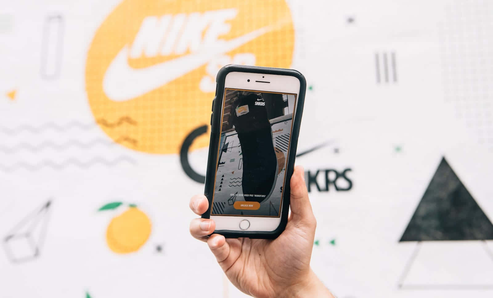 潮流玩家 桌布 壁紙 — 一隻手拿著智慧型手機，顯示一雙黑色Nike運動鞋，背景是充滿活力的幾何圖案牆壁，帶有橙色和黑色圖形。