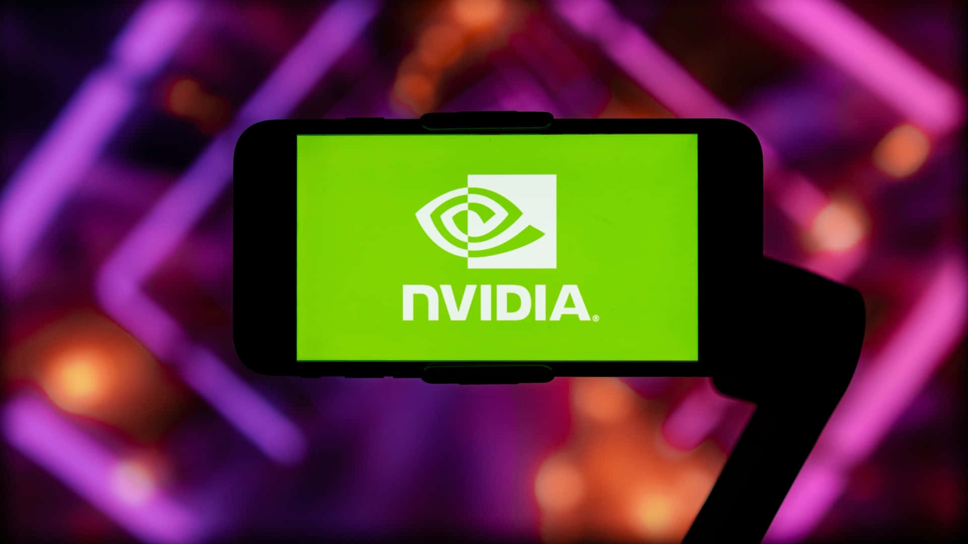 nvidia 桌布 壁紙 — 一隻手拿著智慧型手機，顯示亮綠色NVIDIA標誌，背景是模糊的紫色和橙色霓虹燈。