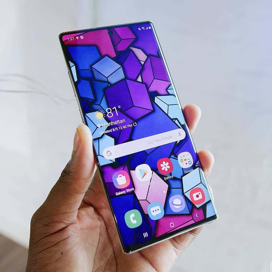 Note 10 桌布 壁紙 — 一隻手拿著智慧型手機，顯示充滿活力的紫色和藍色幾何桌布，帶有應用程式圖示和天氣資訊。