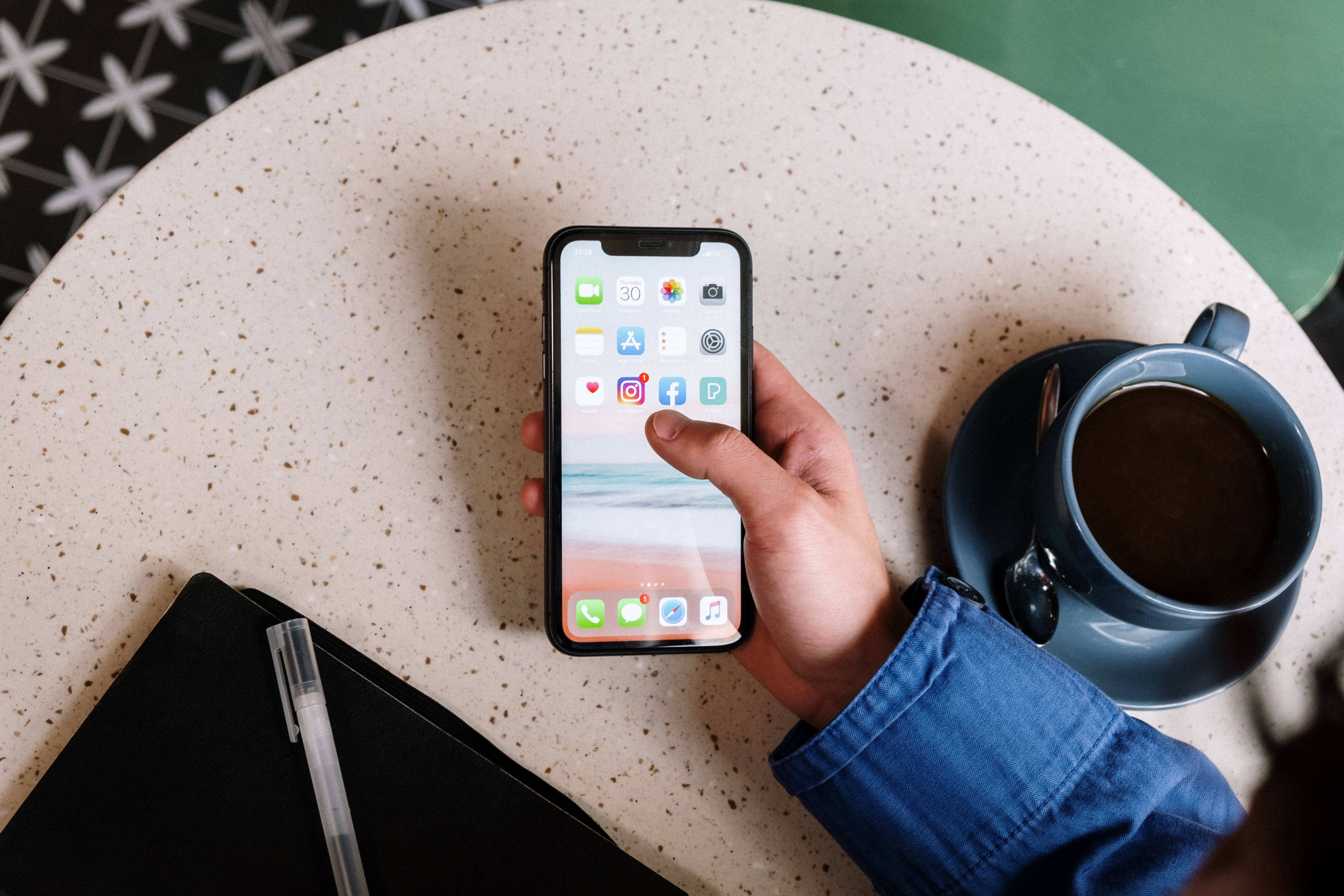 酷炫iPhone 桌布 壁紙 — 一隻手拿著智慧型手機，顯示應用程式圖標，放在有斑點的桌子上，旁邊有咖啡杯和筆記本。
