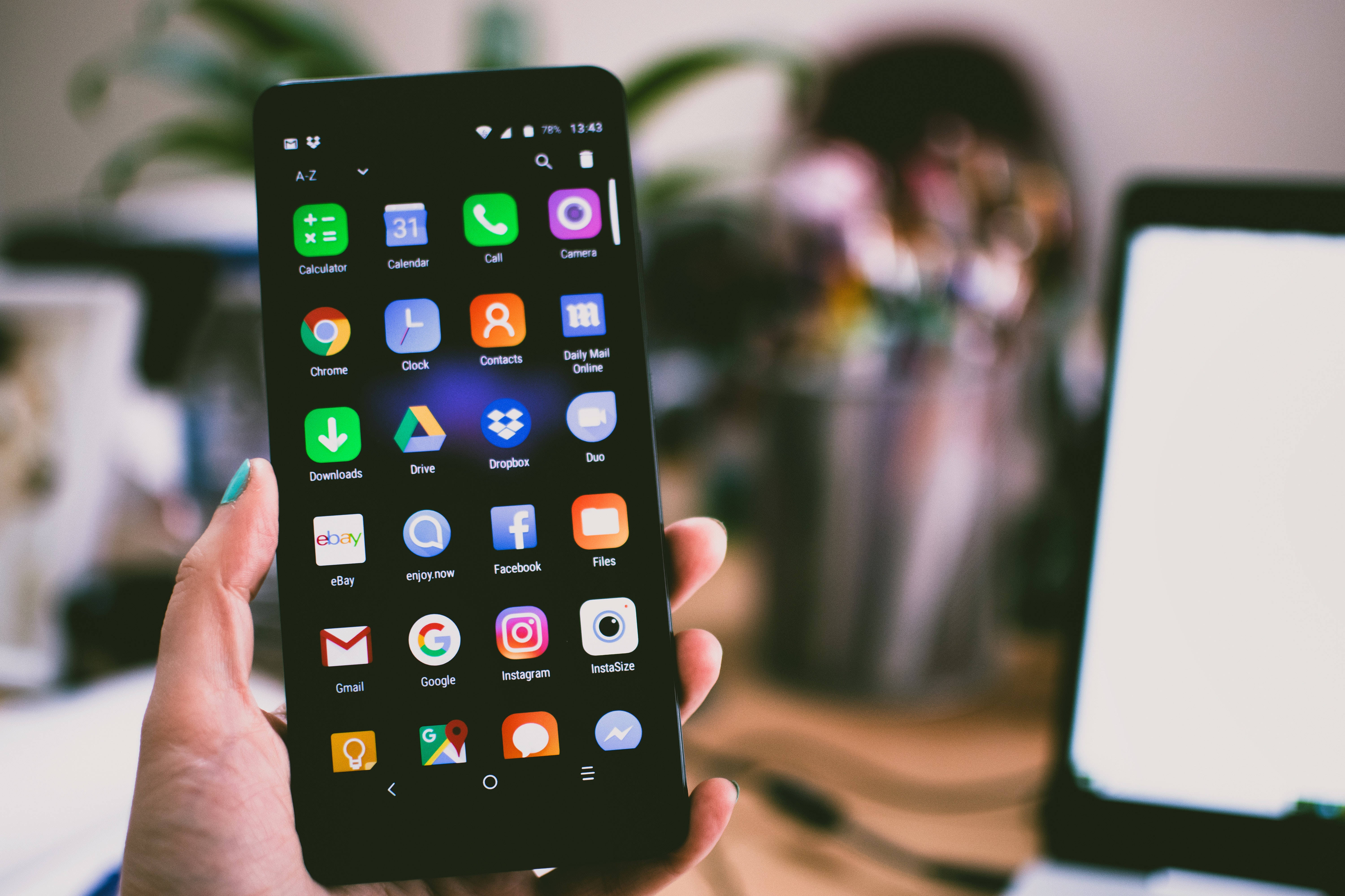 酷炫iPhone 桌布 壁紙 — 一隻手拿著智慧型手機，顯示色彩繽紛的應用程式圖示，背景是模糊的家庭辦公室，有筆記型電腦和植物。