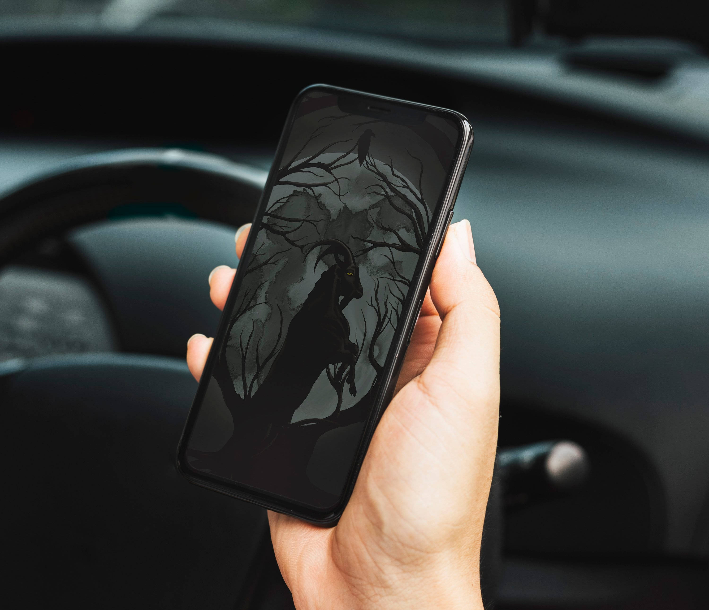 Gothic Phone 桌布 壁紙 — 一隻手拿著智慧型手機，顯示車內扭曲樹木中一個山羊頭人物的黑暗哥德式插圖。