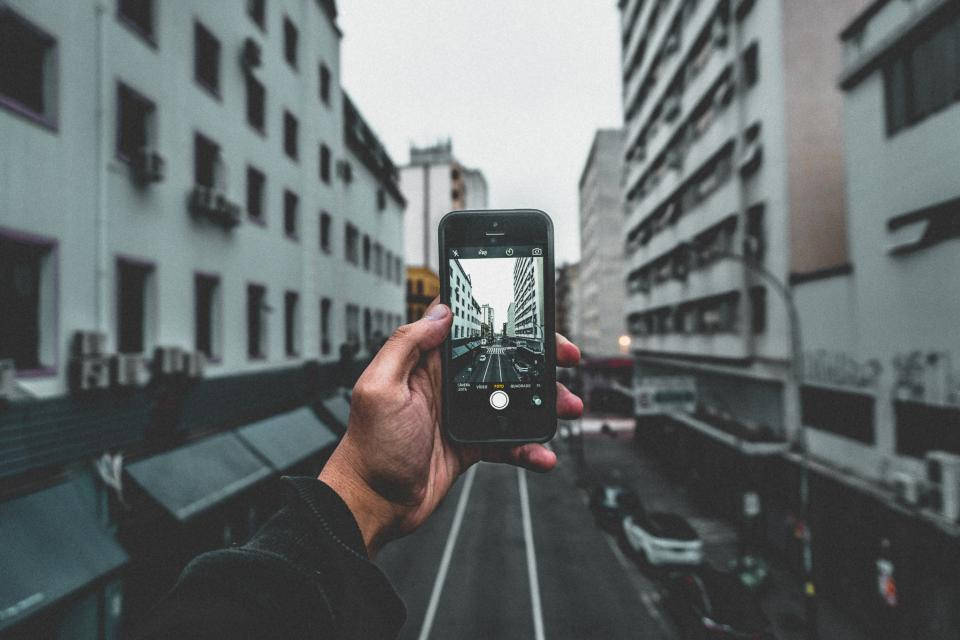 photography iPhone 桌布 壁紙 — 一隻手拿著智慧型手機拍攝一條狹窄的城市街道，兩旁是高樓大廈，天空多雲。
