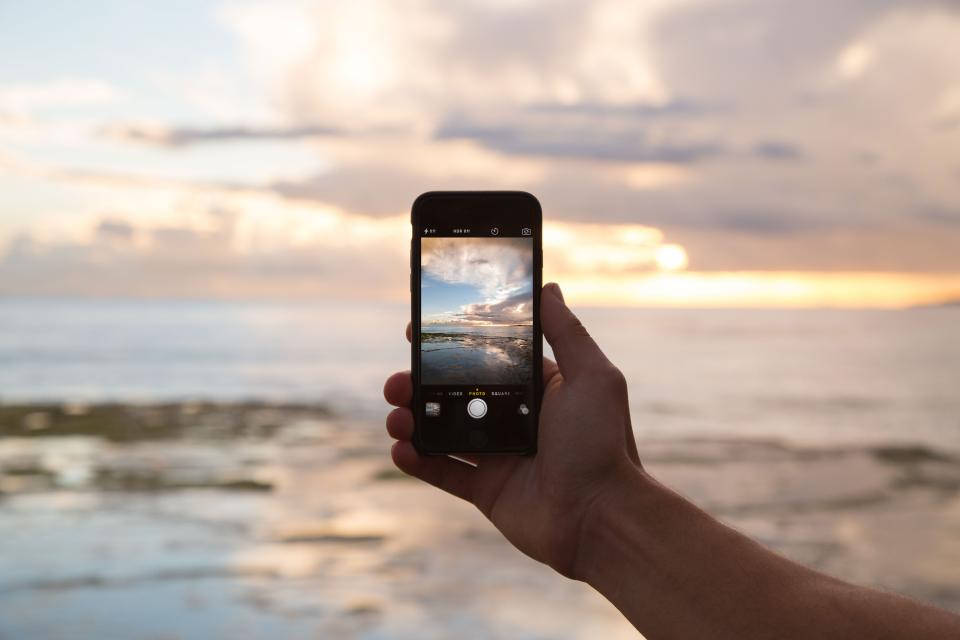 photography iPhone 桌布 壁紙 — 一隻手拿著智慧型手機捕捉海洋上寧靜的日落，伴隨著金色的雲朵和平靜的海水。