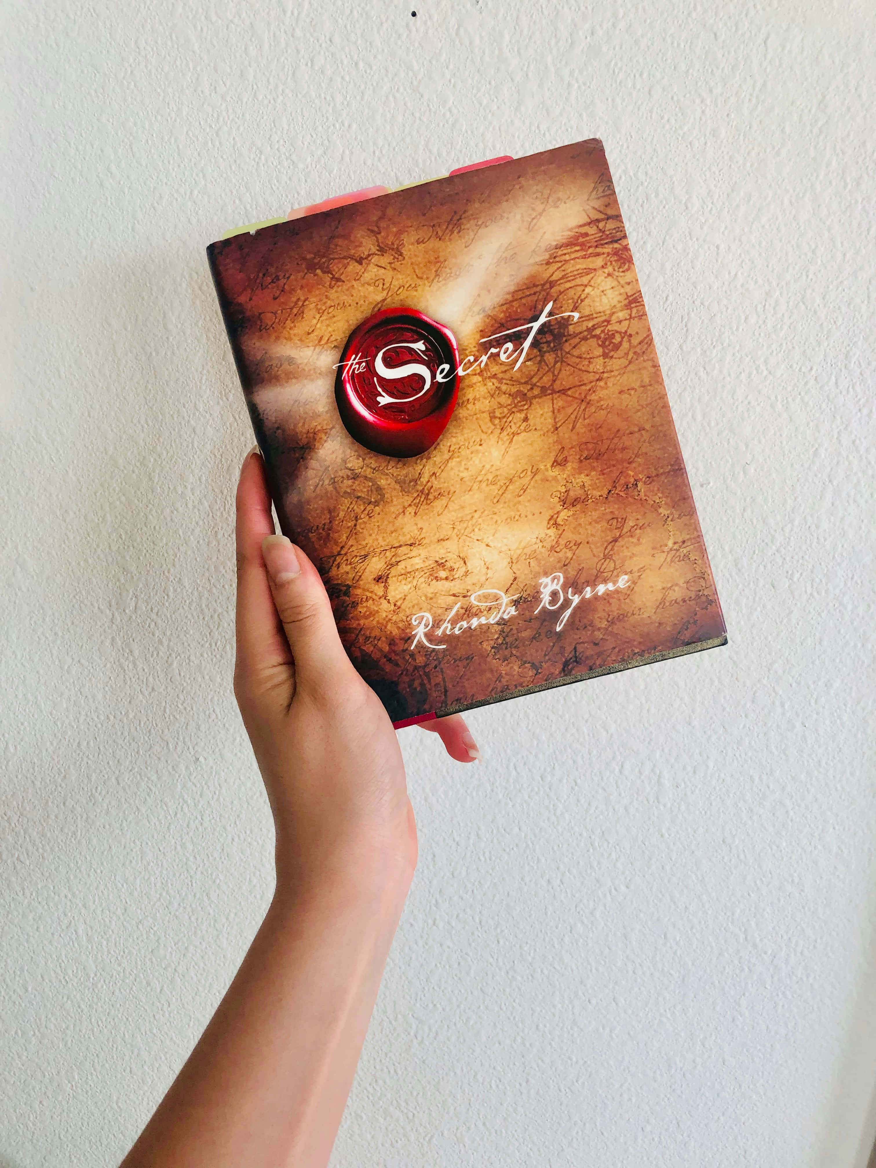 書本美學 桌布 壁紙 — 一隻手拿著朗達·拜恩的《秘密》一書，靠著純白牆壁，書本有紋理封面和紅色火漆印章。