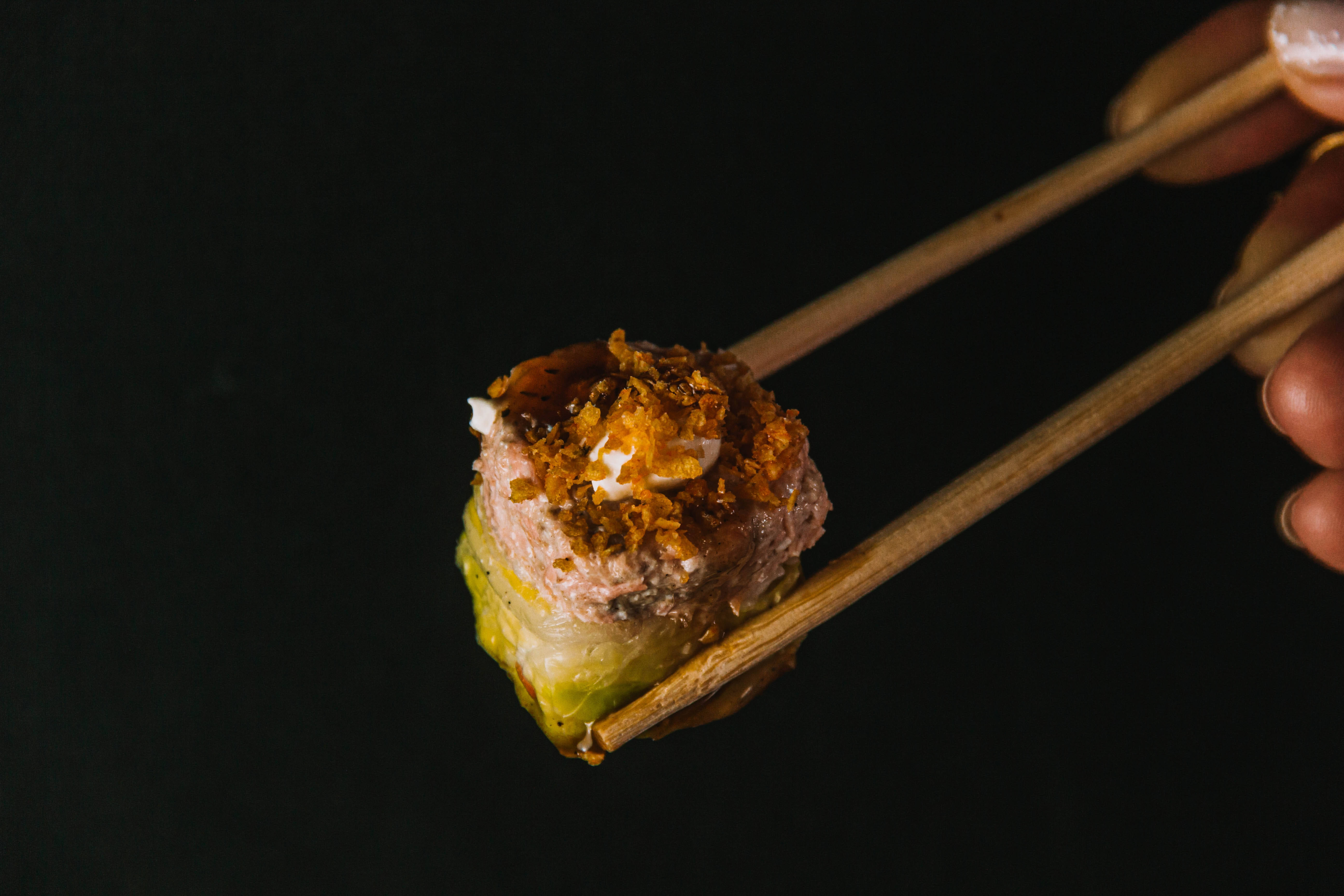 Sushi 桌布 壁紙 — 一隻手拿著木筷子，夾起一個閃亮的壽司卷，上面鋪滿酥脆的金色薄片，背景是純黑色。