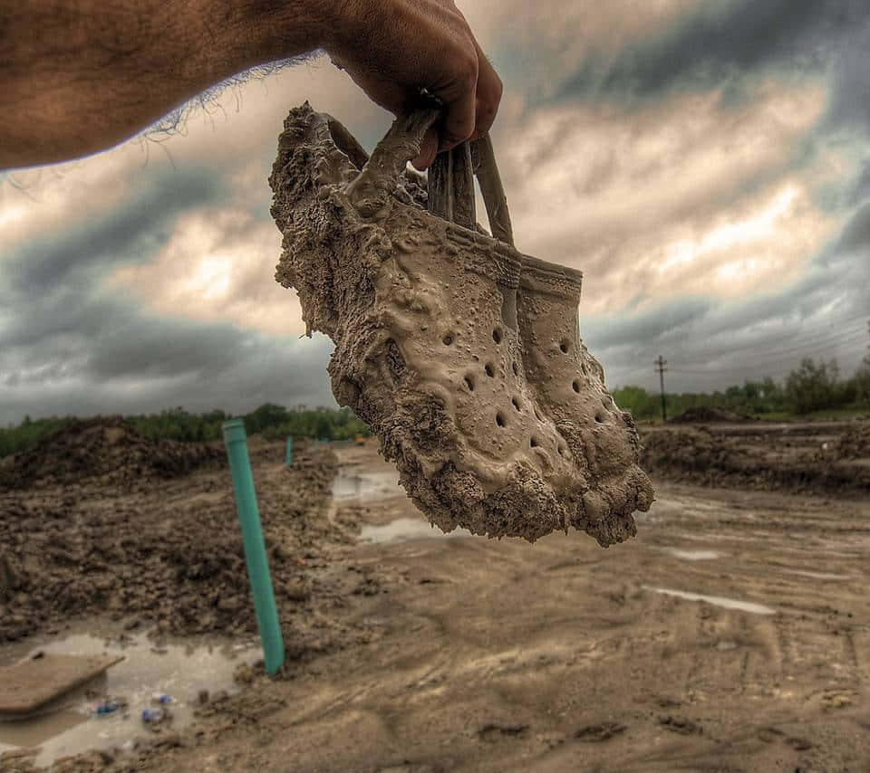 Crocs 桌布 壁紙 — 一隻手拿著沾滿泥巴的 Croc 涼鞋，背景是陰沉多雲的天空，下方是淹水的泥濘田野。