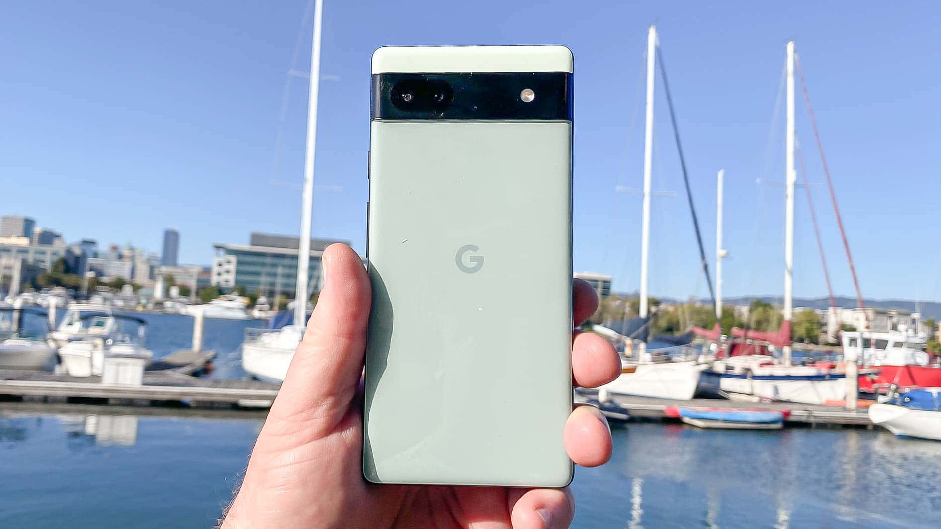 google 桌布 壁紙 — 一隻手拿著淺綠色 Google Pixel 手機，背景是帆船和城市天際線，在晴朗的藍天下。