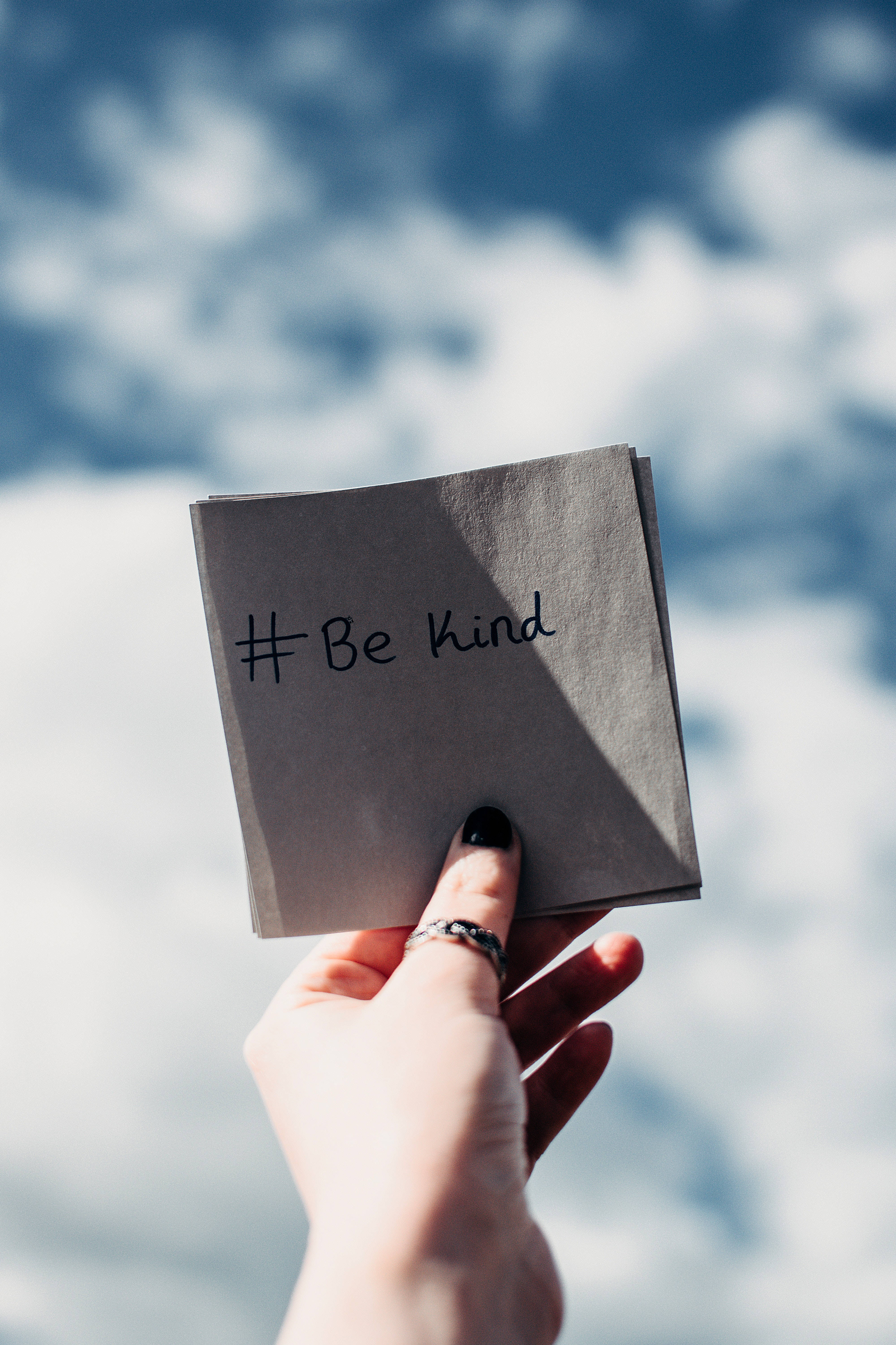 手機 桌布 壁紙 — 一隻手拿著灰色餐巾，上面有手寫的 hashtag be kind，背景是明亮的藍天和 fluffy 的白雲。