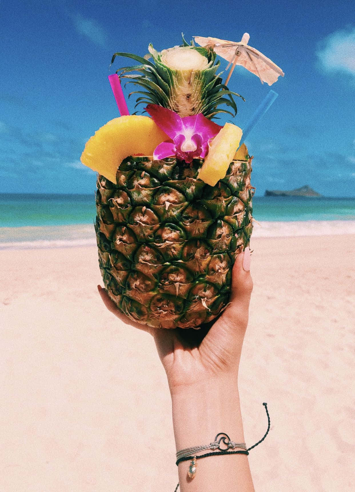 Pineapple 桌布 壁紙 — 一隻手拿著熱帶鳳梨飲品，飾以水果和花朵，背景是明亮的沙灘。