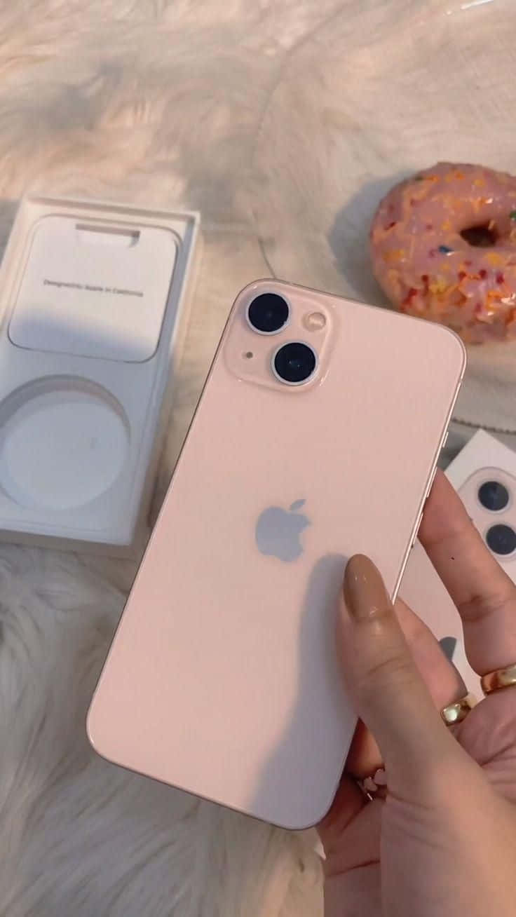 iPhone 12 桌布 壁紙 — 一隻手拿著玫瑰金色 iPhone 13 mini，配以甜甜圈和 MagSafe 充電器，放在毛絨白色表面上。