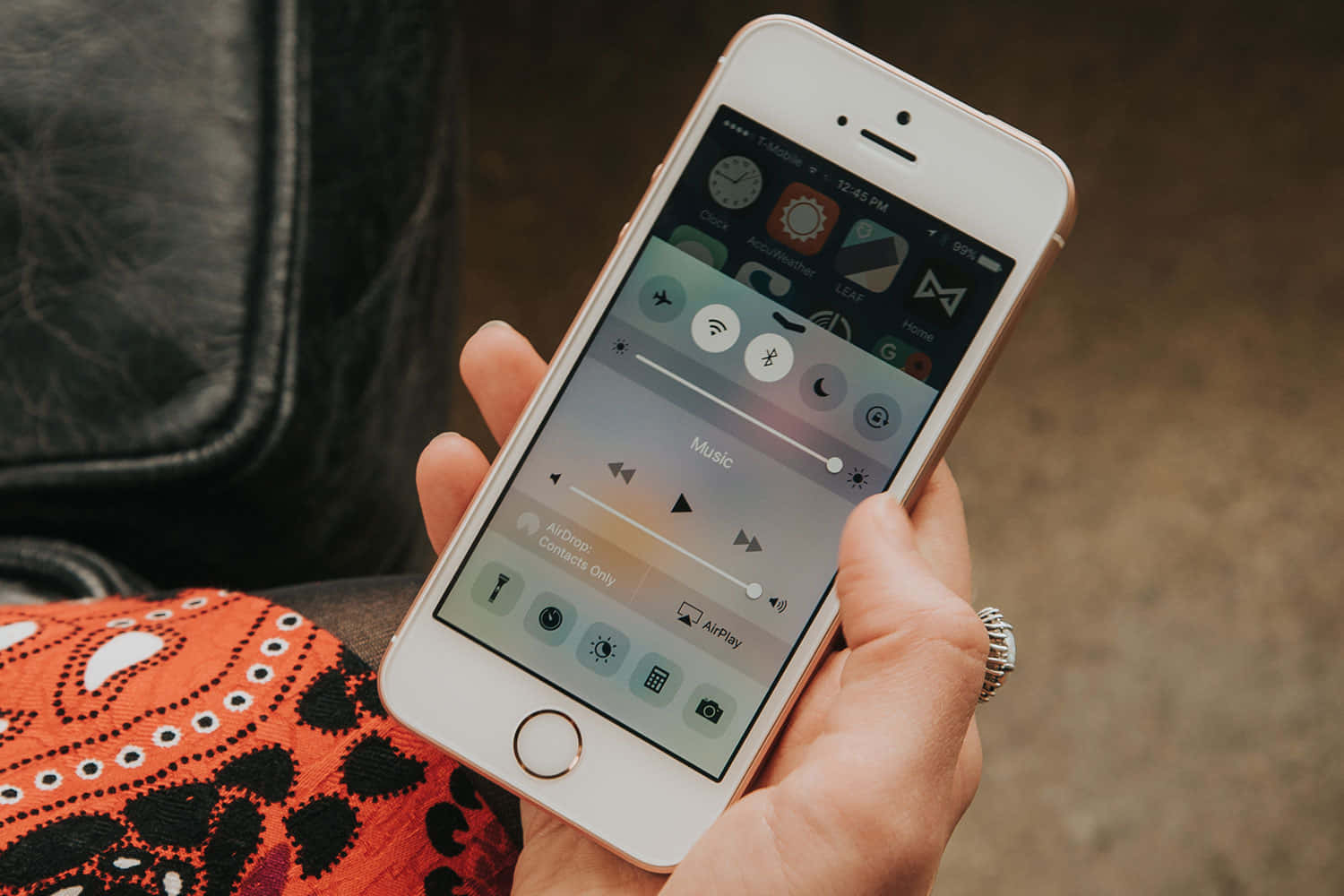 Apple Phone 桌布 壁紙 — 一隻手拿著玫瑰金色iPhone，顯示控制中心，包含音樂播放控制和應用程式圖示，背景為橙色圖案布料。