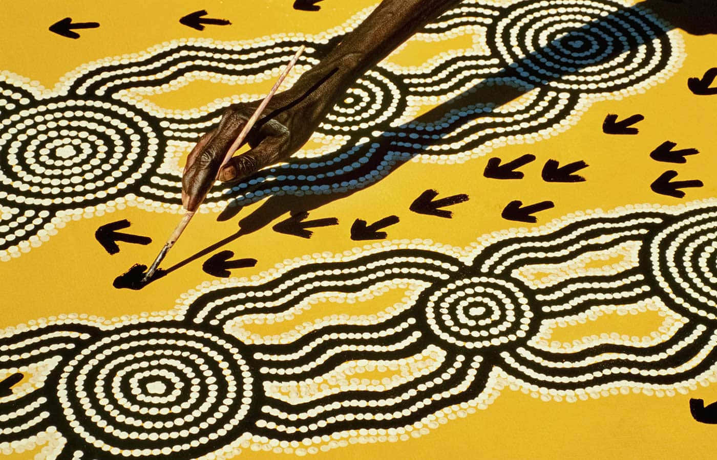 Aboriginal 桌布 壁紙 — 一隻手拿著畫筆在明亮的陽光下，在充滿活力的黃色畫布上創作出複雜的點狀圖案和箭頭。