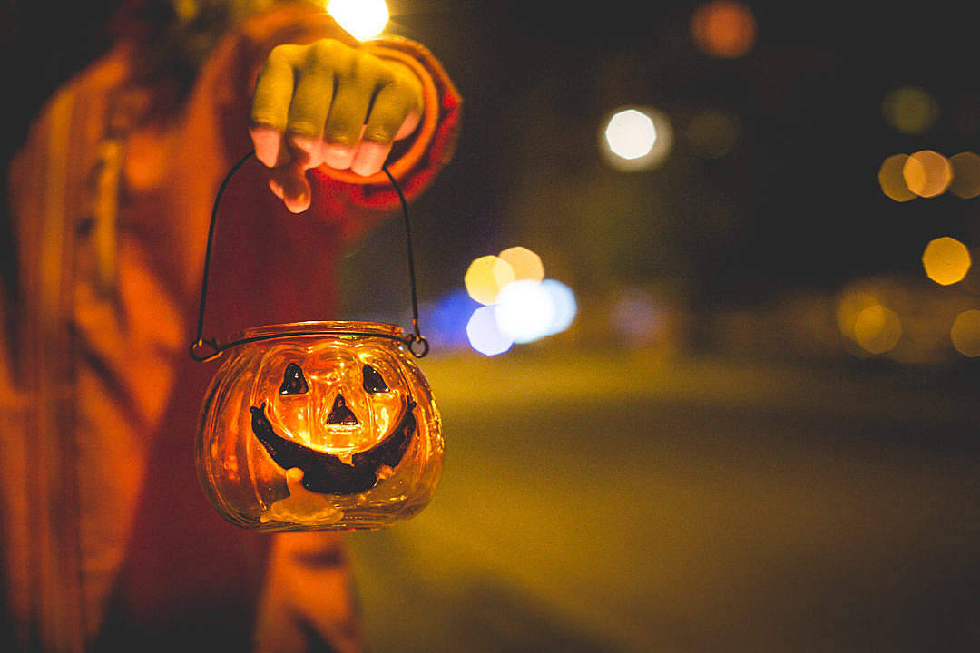 Cute Halloween Desktop 桌布 壁紙 — 一隻手拿著發光的傑克南瓜燈籠，背景是模糊的夜晚街道和散景燈光。
