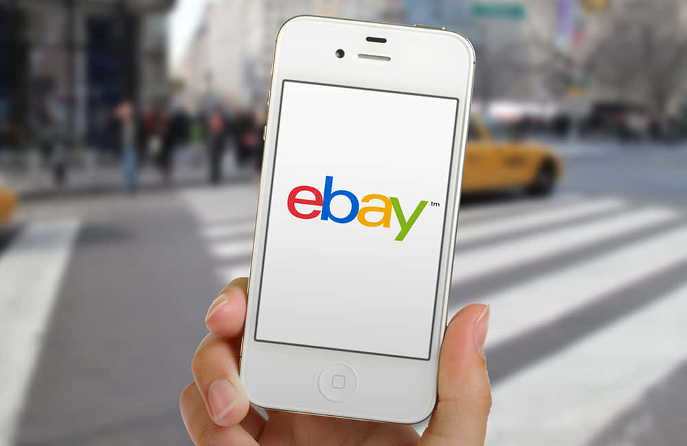品牌 桌布 壁紙 — 一隻手拿著白色iPhone，顯示色彩繽紛的eBay標誌，背景是模糊的城市街道和一輛黃色的士。