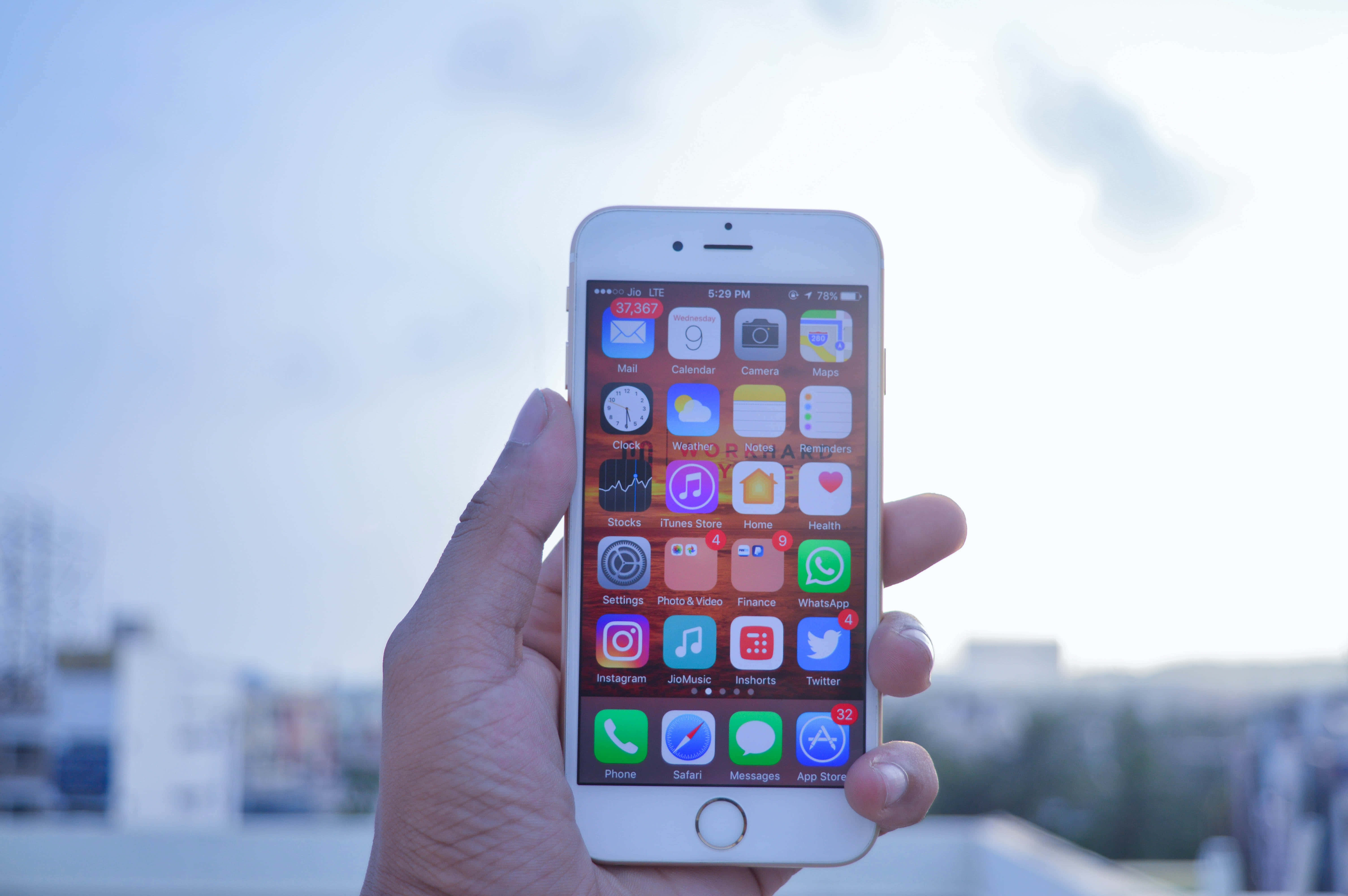 Apple Phone 桌布 壁紙 — 一隻手拿著白色iPhone，顯示色彩繽紛的應用程式圖標，背景是明亮朦朧的天空和模糊的城市建築。