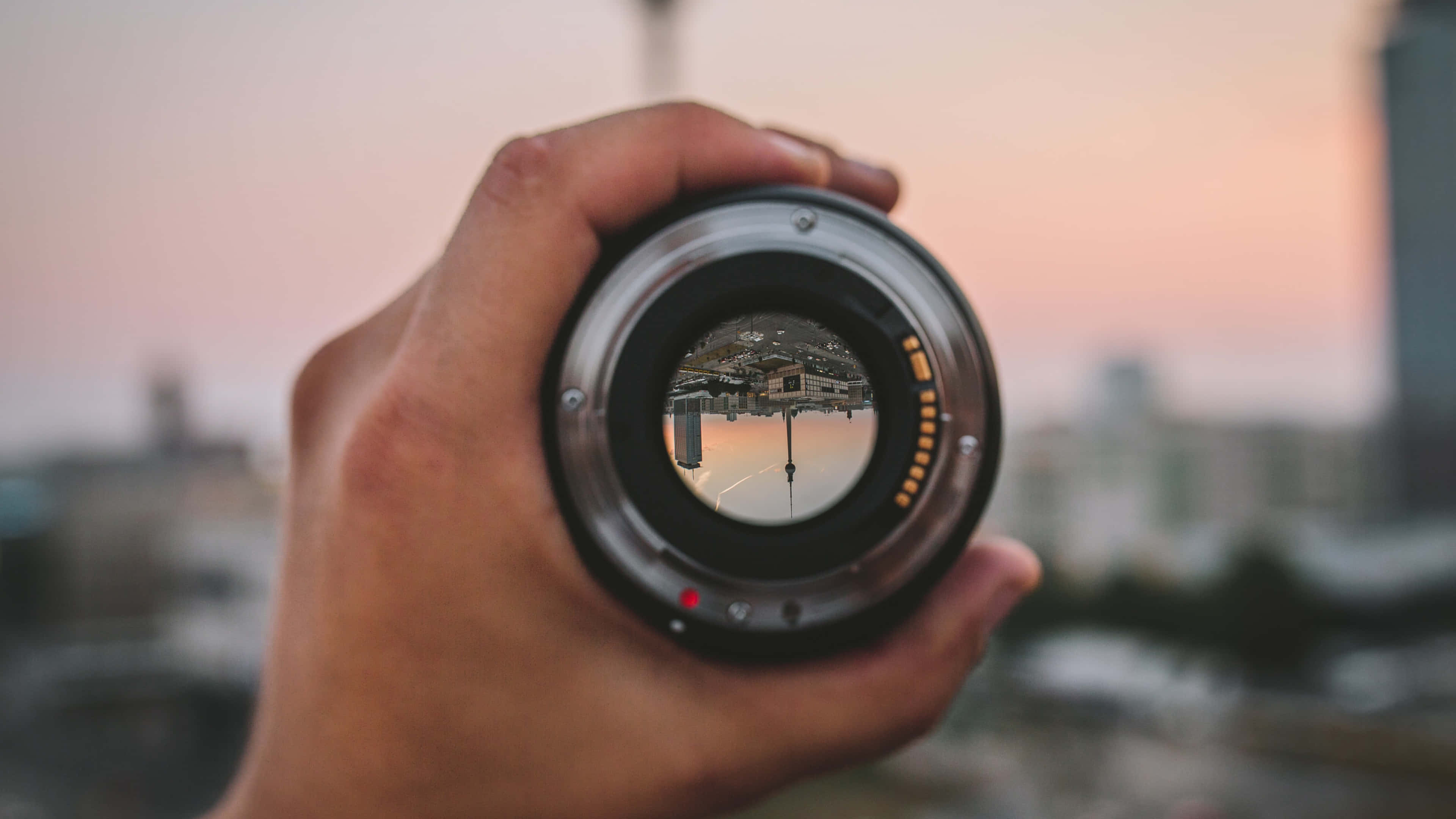 Dslr 桌布 壁紙 — 一隻手拿著相機鏡頭，反射出日落時分溫暖粉紅和橙色調的城市景觀。