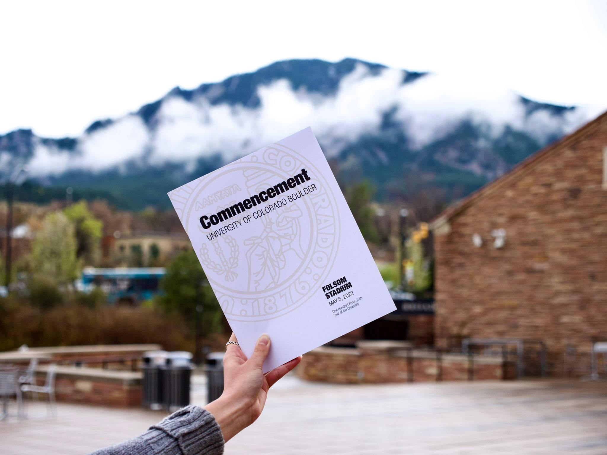 Colorado 桌布 壁紙 — 一隻手拿著科羅拉多大學博爾德分校的畢業典禮程序單，背景是霧濛濛的山脈和校園的磚造建築。