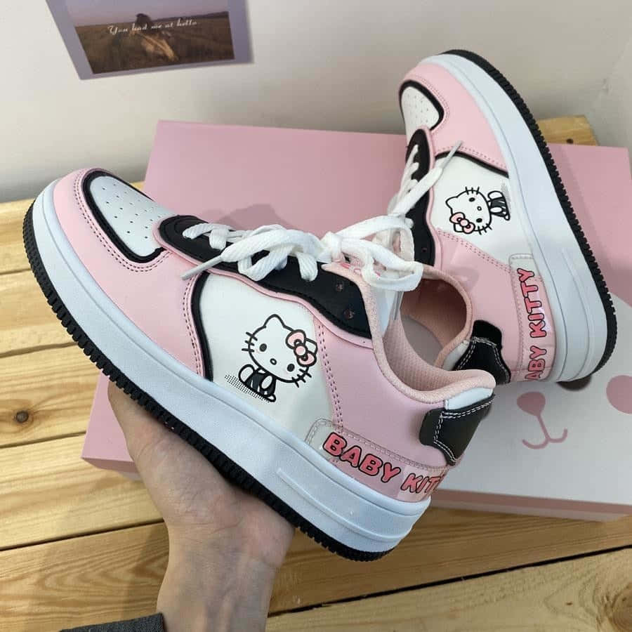 Kawaii Hello Kitty 桌布 壁紙 — 一隻手拿著粉紅、黑、白色的 Baby Kitty 運動鞋，上面有 Hello Kitty 圖案，背景是木質表面和粉紅色盒子。
