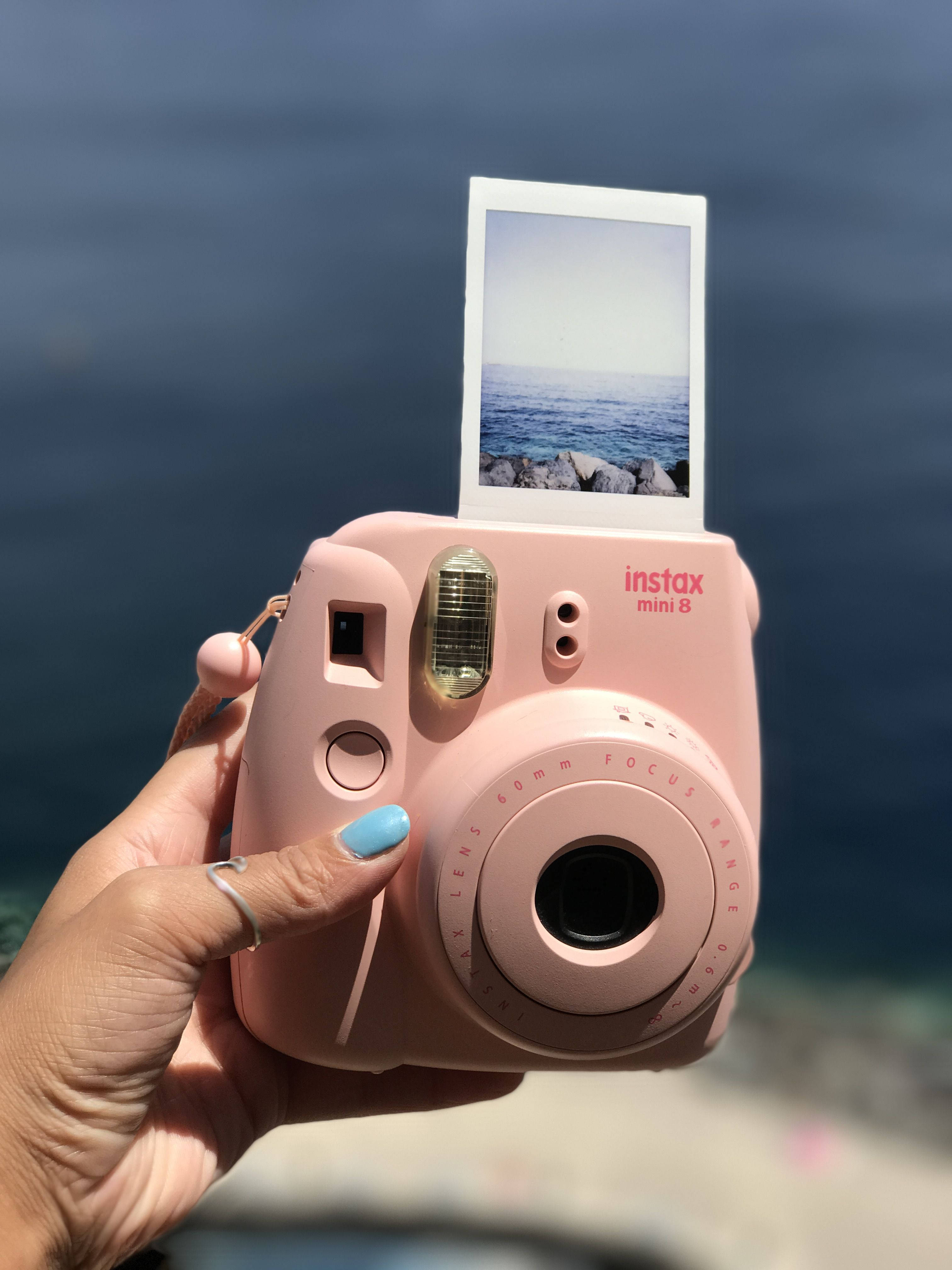 相機 桌布 壁紙 — 一隻手拿著粉紅色Instax Mini 8相機，一張列印的海洋照片在模糊的藍色海洋背景前顯現。