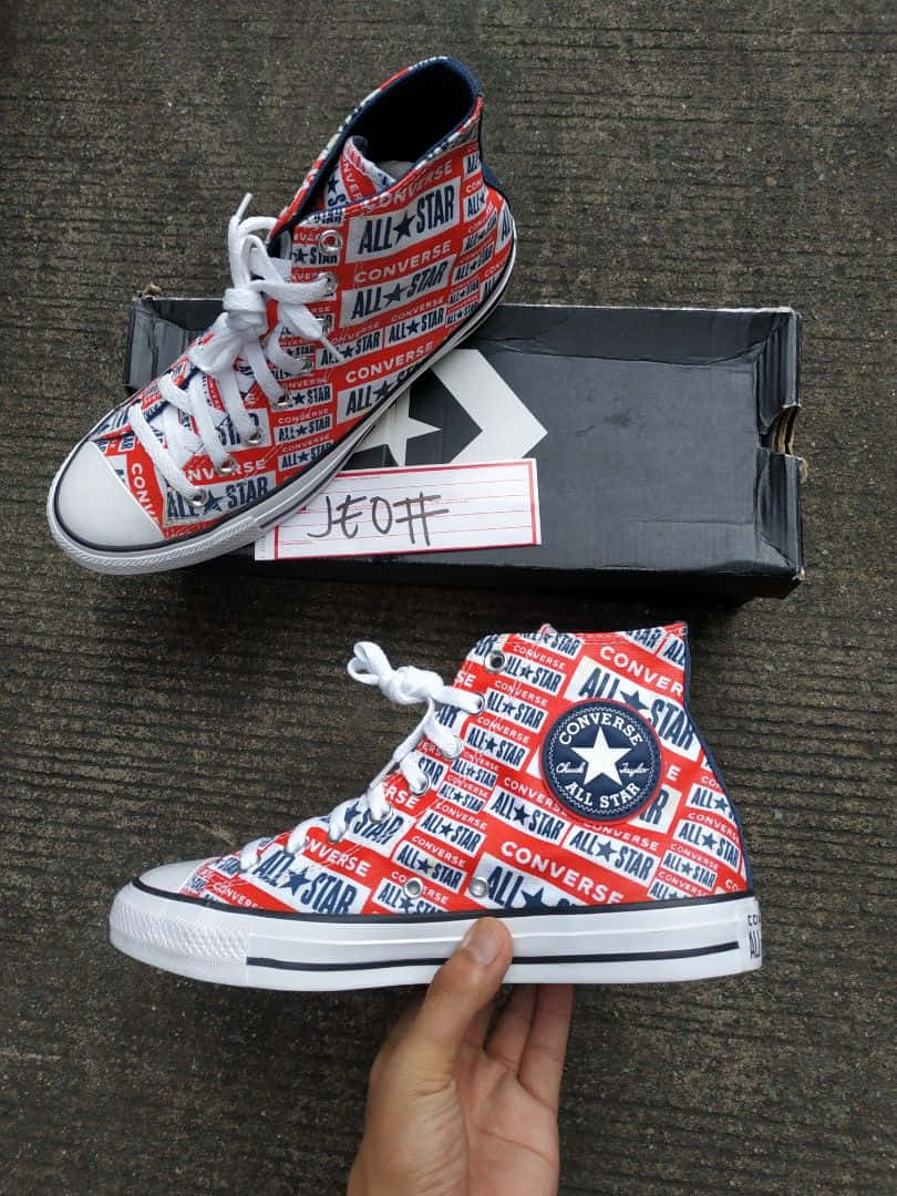 Converse 桌布 壁紙 — 一隻手拿著紅白藍色的Converse All Star高筒運動鞋，鞋上重複印有品牌標誌，背景是紋理灰色地板。