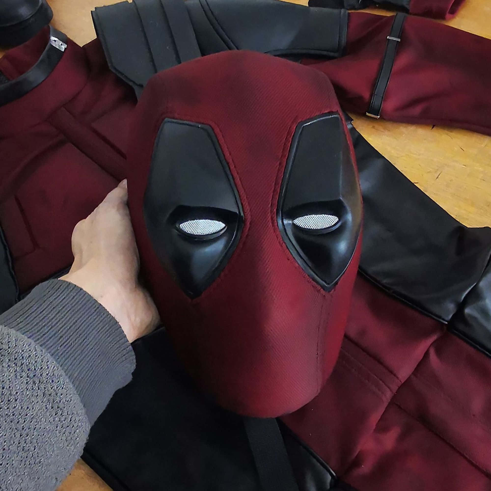 Deadpool Mask 桌布 壁紙 — 一隻手拿著紅黑色的死侍面具，放在木地板上鋪開的配套服裝上方。