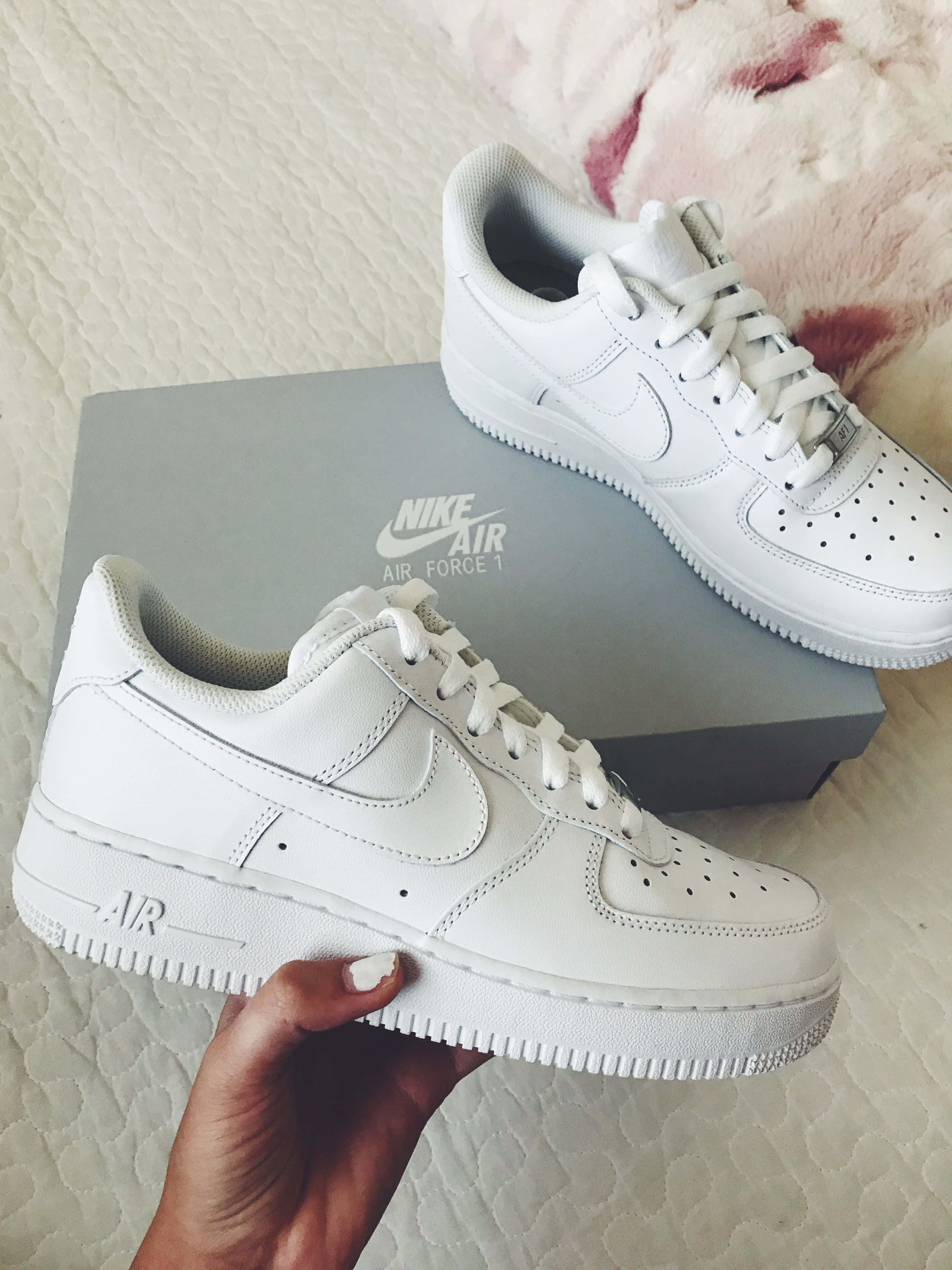 Nike Air Force 桌布 壁紙 — 一隻手拿著純白色的Nike Air Force 1運動鞋，旁邊放著灰色鞋盒，置於有紋理的白色表面上。