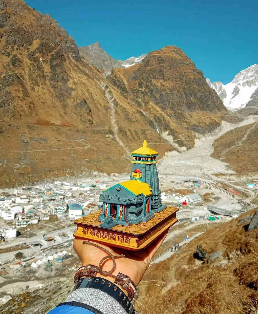 Kedarnath 桌布 壁紙 — 一隻手拿著色彩繽紛的凱達爾納特神廟微型模型，背景是崎嶇的喜馬拉雅山脈和廣闊的山谷小鎮，天空清澈湛藍。