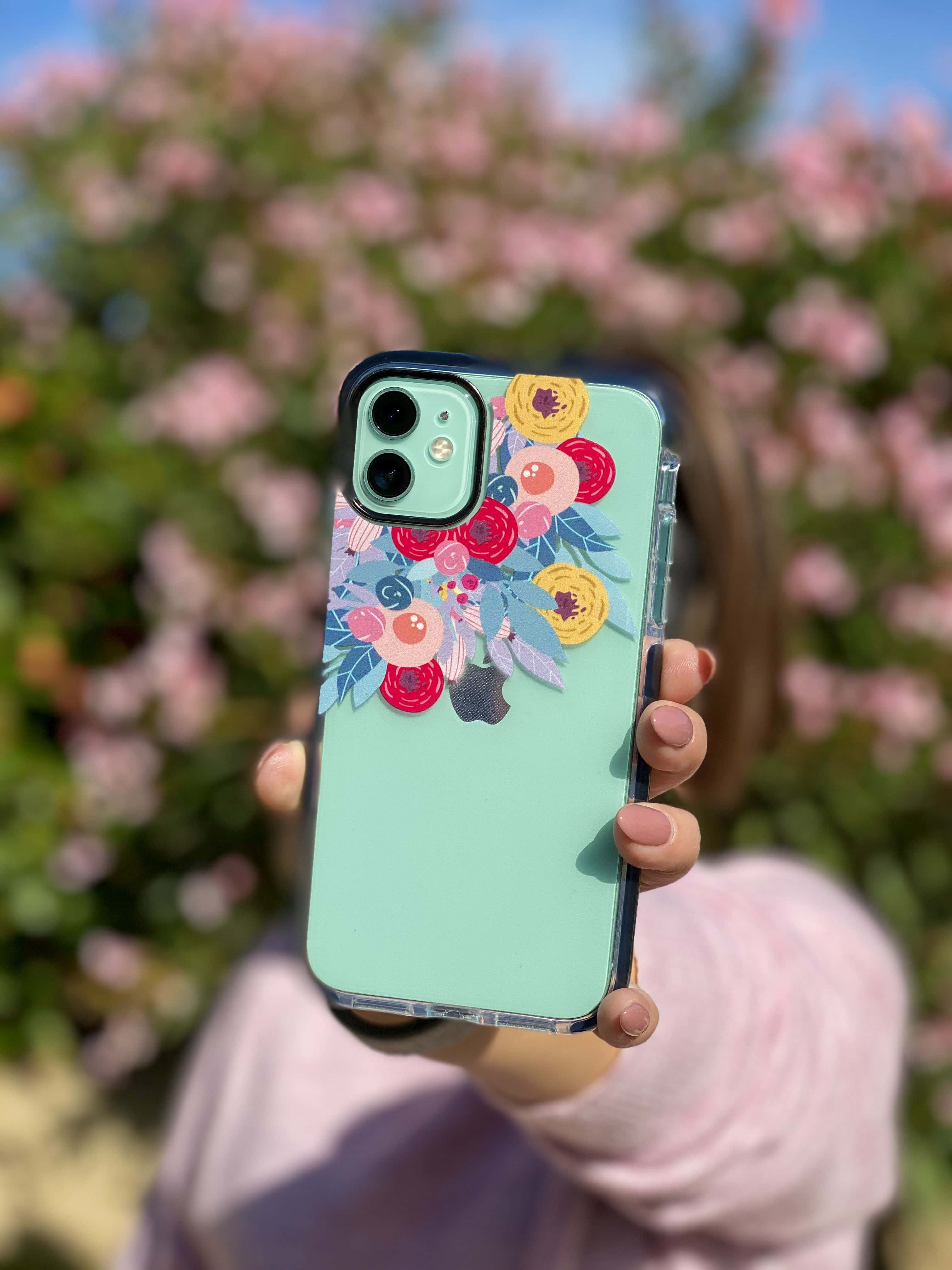 Phone Case 桌布 壁紙 — 一隻手拿著薄荷綠色手機殼，上面有彩色花卉貼紙，背景是粉紅花朵和藍天。