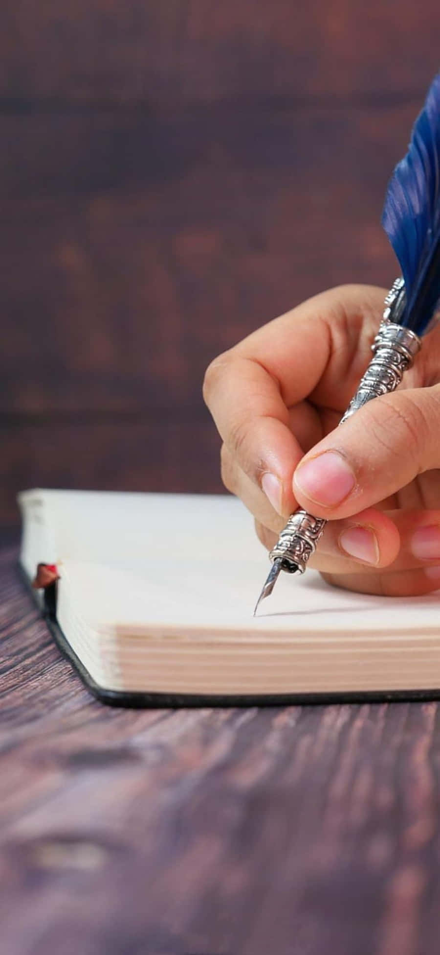 Pen 桌布 壁紙 — 一隻手拿著藍色羽毛筆，懸停在放在深色木質表面上的空白筆記本上方。