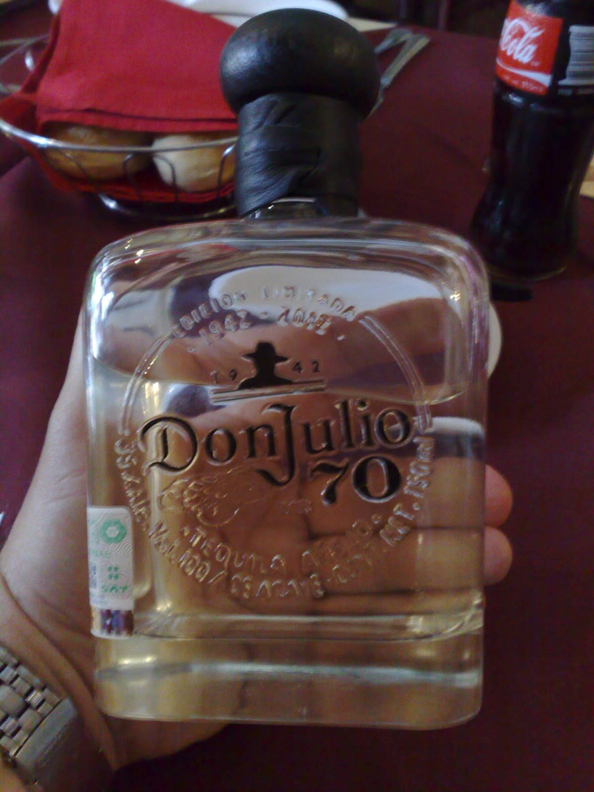 glass 桌布 壁紙 — 一隻手拿著透明的Don Julio 70龍舌蘭酒瓶，黑色瓶蓋，紅色桌布上，背景有可口可樂瓶。