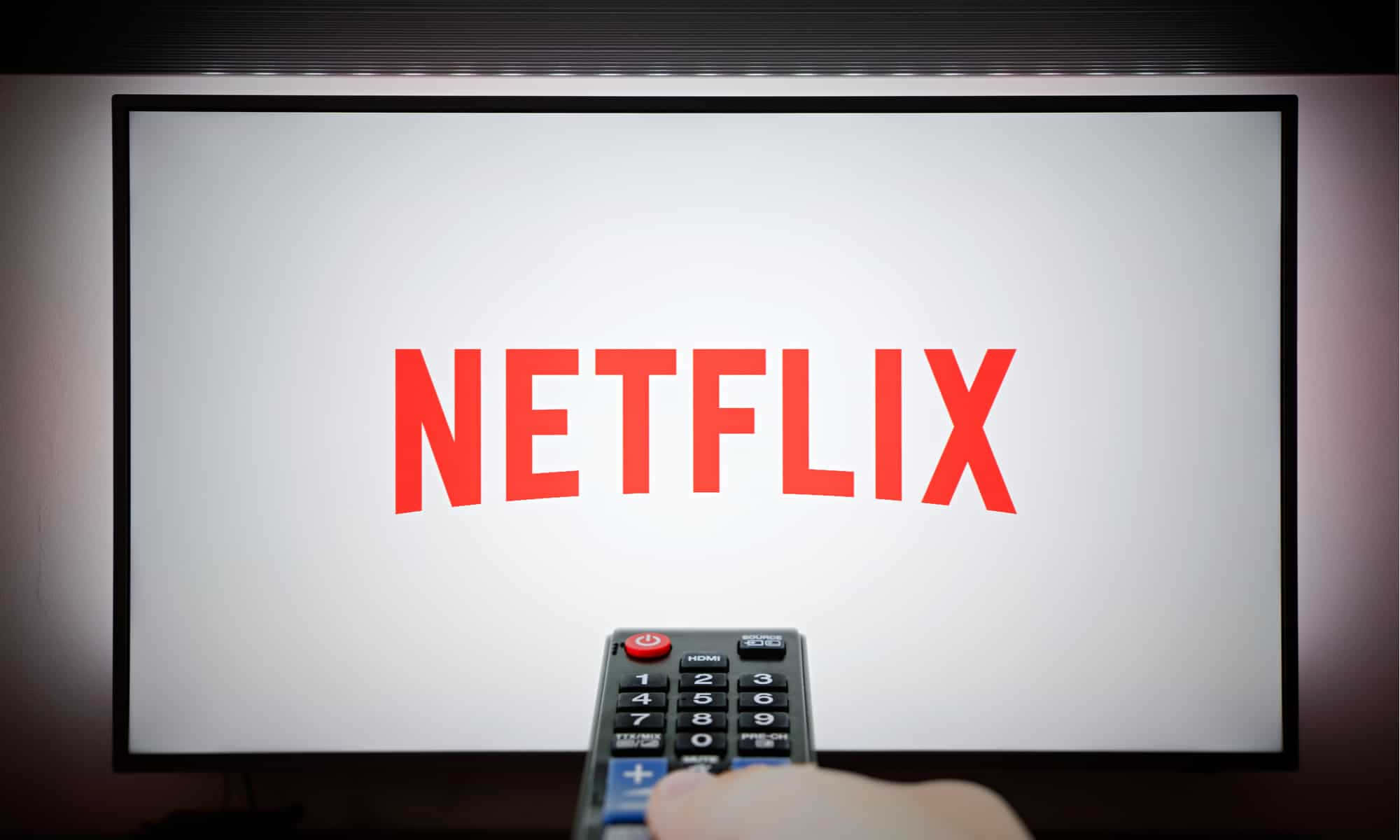 Netflix 桌布 壁紙 — 一隻手拿著遙控器，對準一個顯示紅色Netflix標誌的電視螢幕，背景為亮白色。