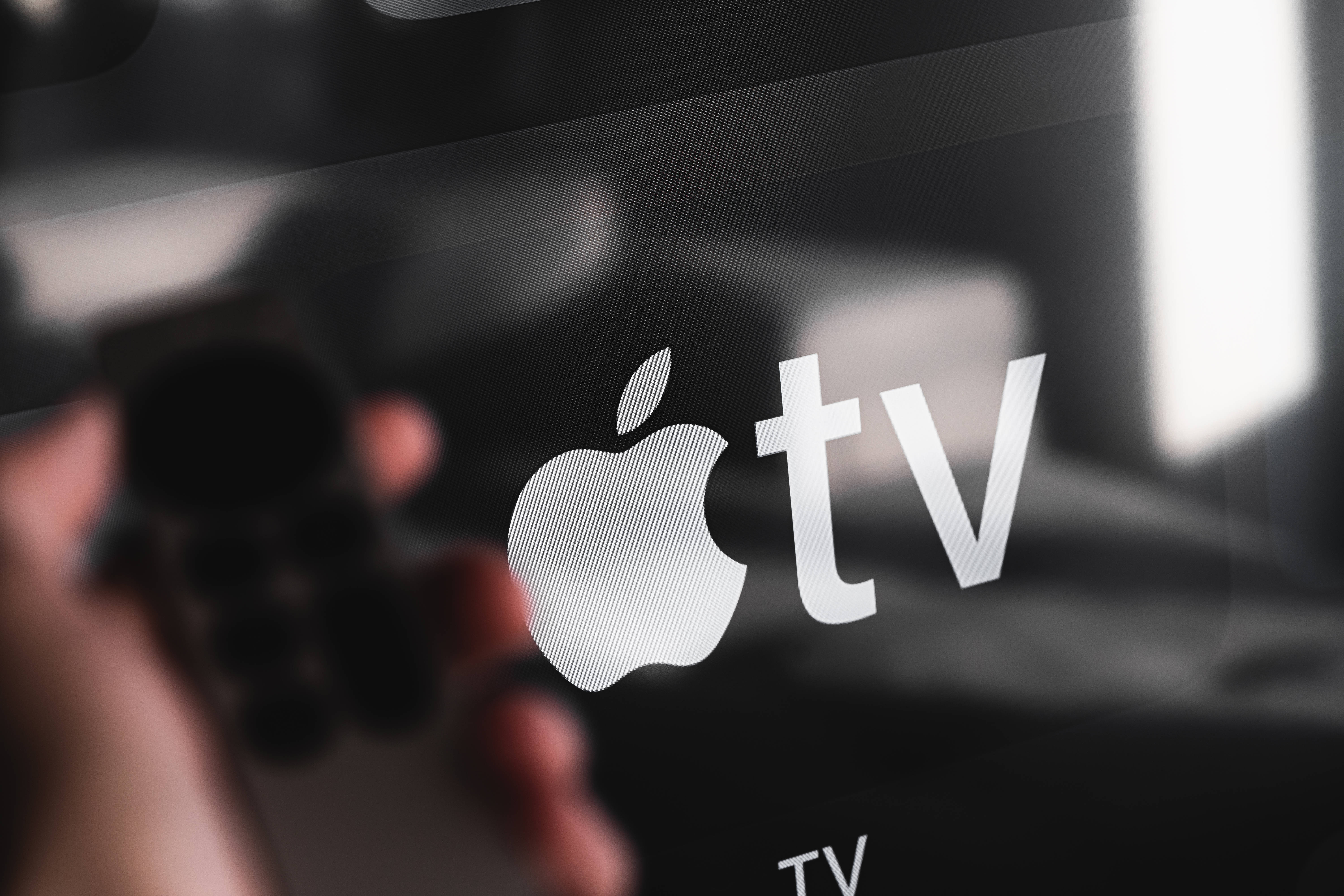 標誌 桌布 壁紙 — 一隻手拿著遙控器，對準發光的Apple TV螢幕，標誌清晰對焦，背景模糊昏暗。