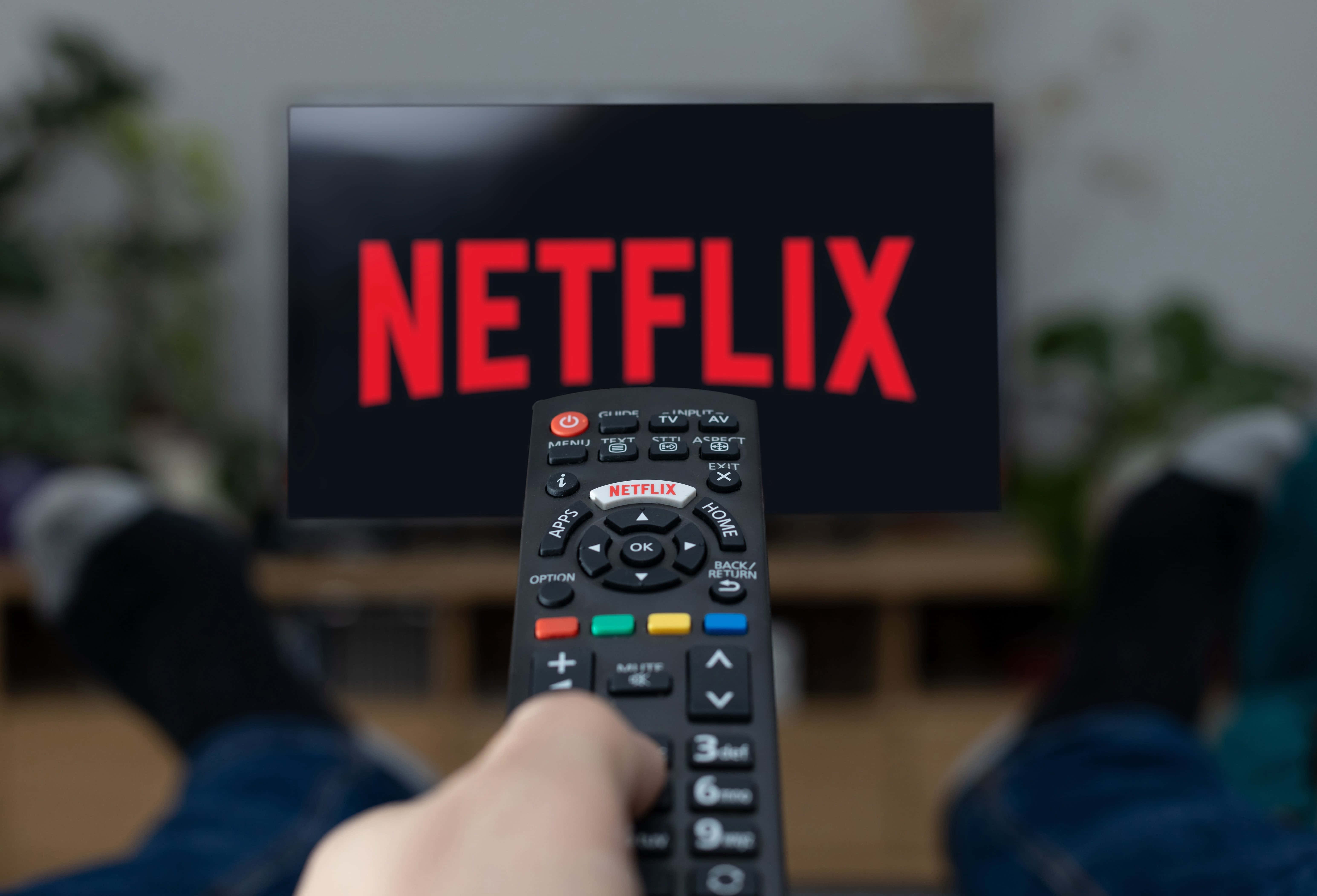 Netflix 桌布 壁紙 — 一隻手拿著遙控器，指向顯示紅色 Netflix 標誌的電視螢幕，在舒適的客廳環境中。
