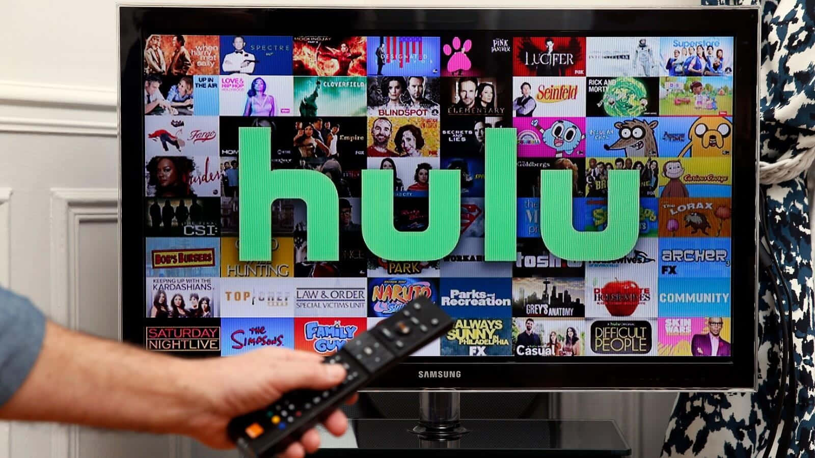 Hulu 桌布 壁紙 — 一隻手拿著遙控器指向電視，電視上顯示Hulu標誌，背景是色彩繽紛的節目縮圖網格。