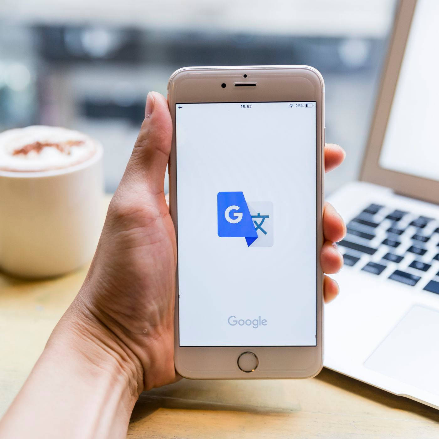 Google Translate 桌布 壁紙 — 一隻手拿著金色iPhone，顯示Google翻譯應用程式，背景有筆記型電腦和咖啡杯，呈現柔和焦點。